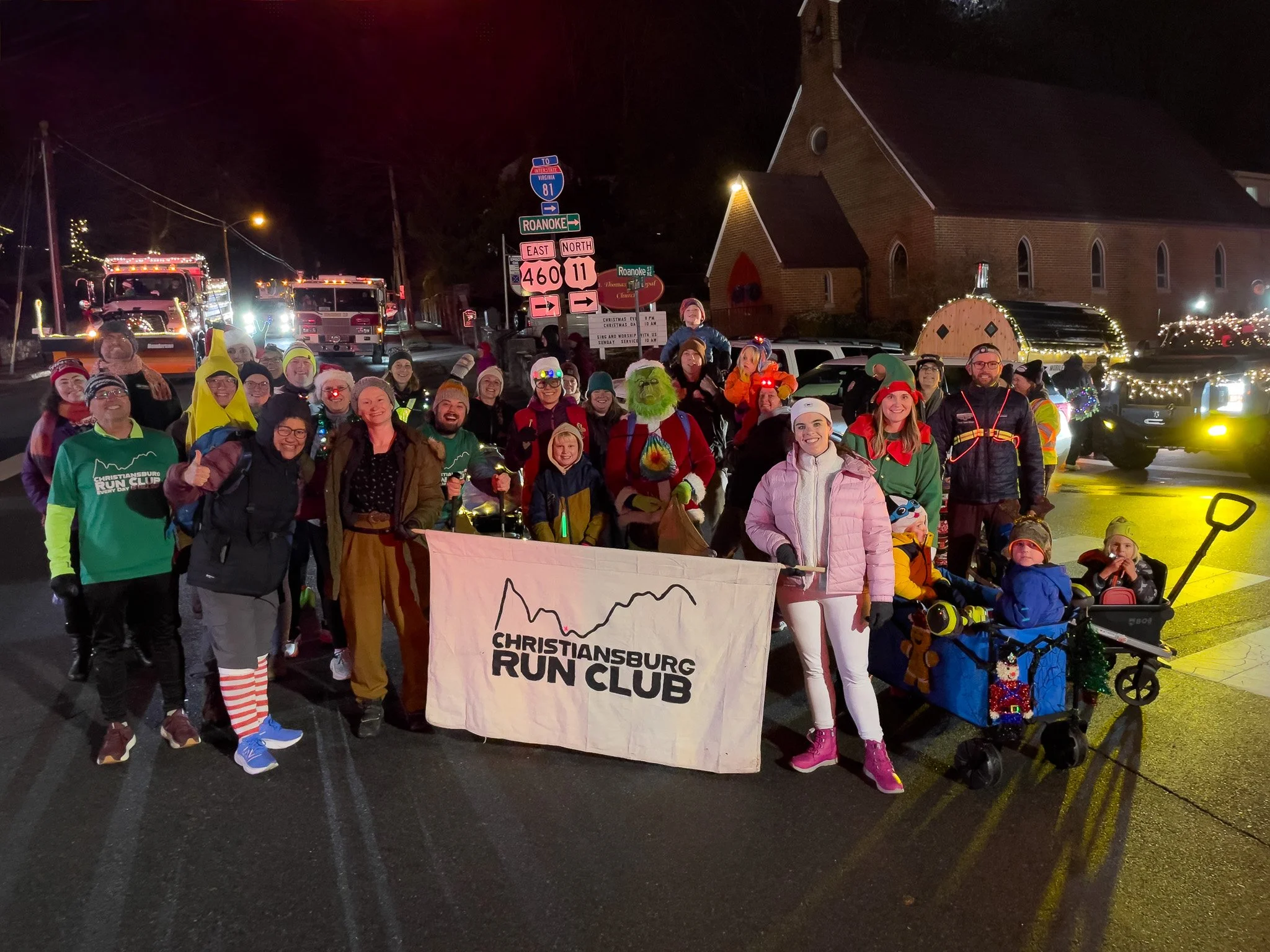 A large group of people dressed in festive and athletic clothing, holding a banner that reads 'Christiansburg Run Club,' gathered on a street at night for a holiday-themed event. Some are wearing Santa hats, elf costumes, and colorful sweaters, with 