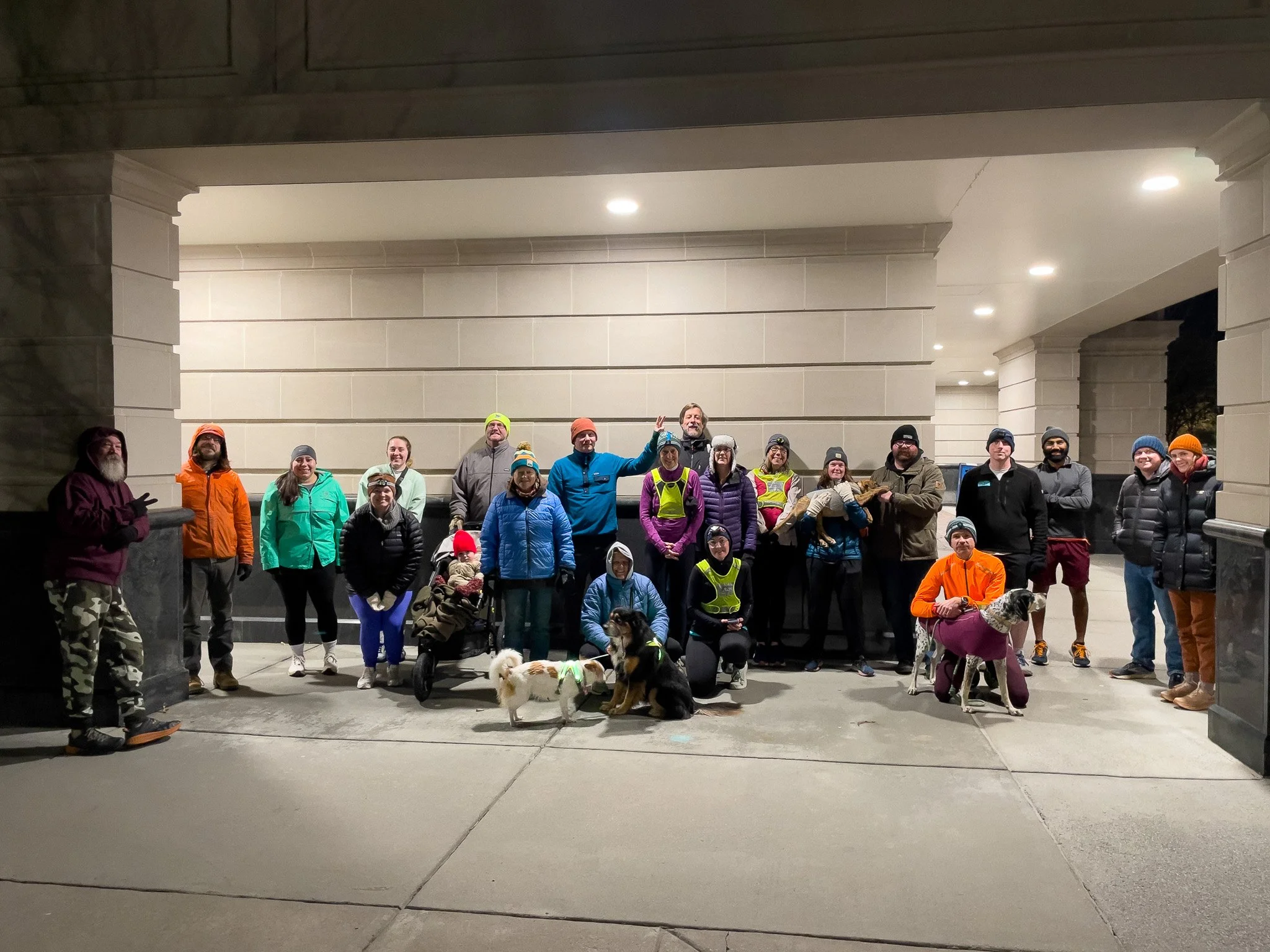 Group of people, some with dogs, gathered under a lit bridge at night, dressed in warm outdoor clothing.
