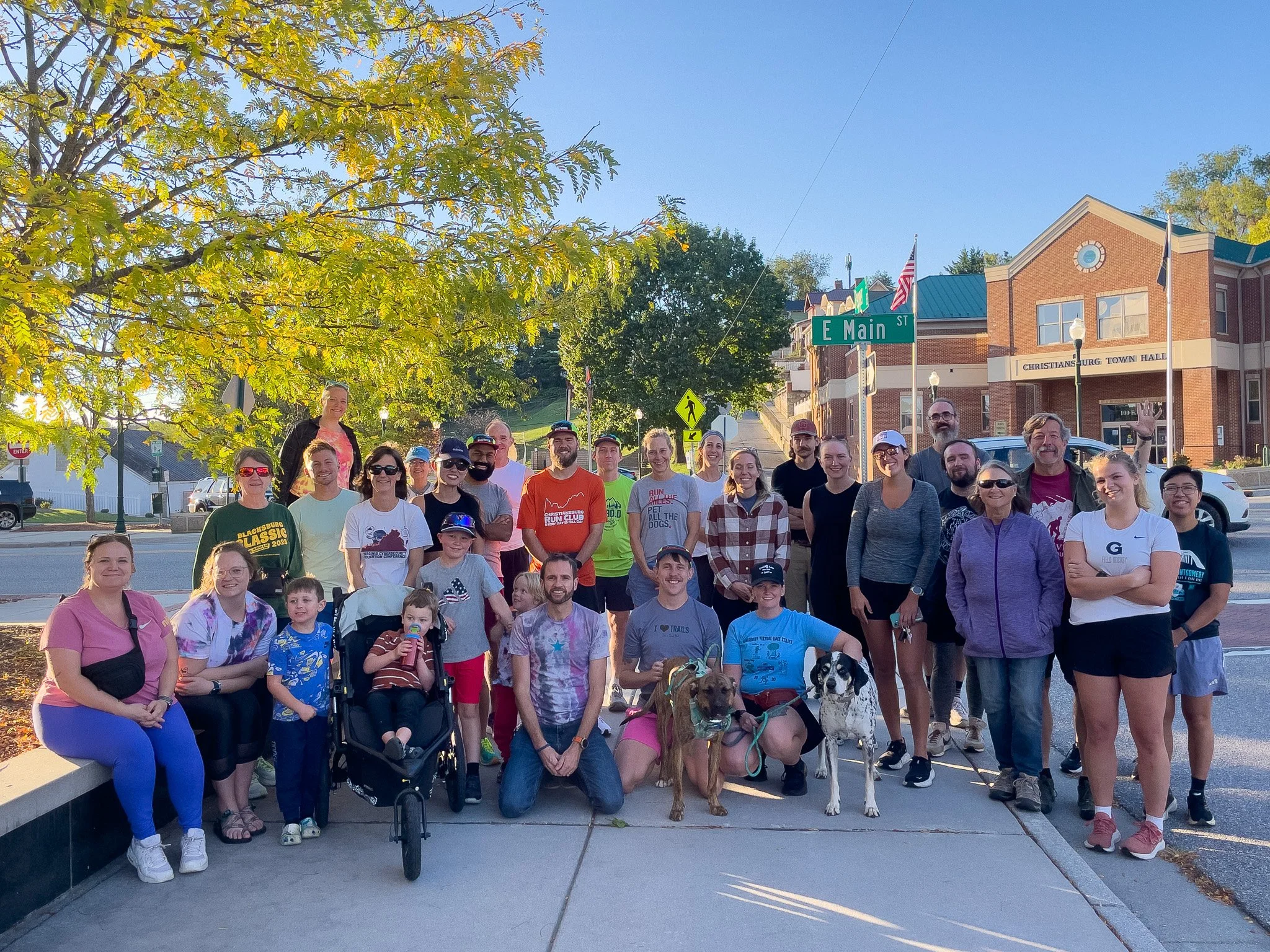 Group of people including children and adults gathered outdoors for a photo in front of a building with signs for E Main Street and Christiansburg Town Hall on a sunny day with green trees in the background.