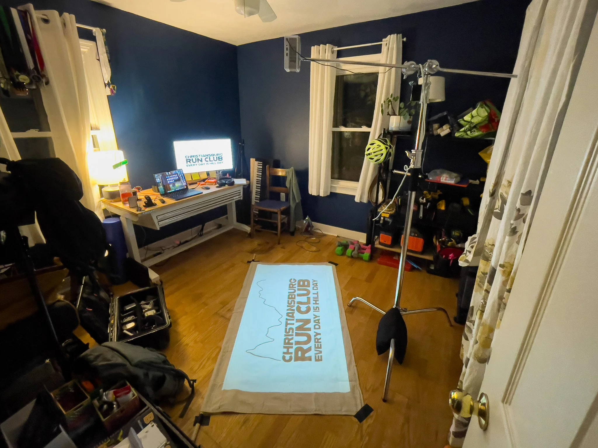 A room set up for a virtual run event, featuring a projection screen on a mat with 'Christiansburg Run Club' and 'Every Day Is Hall Day' displayed, a stationary bike with a helmet, a desk with a monitor, keyboard, and various running gear, a window w