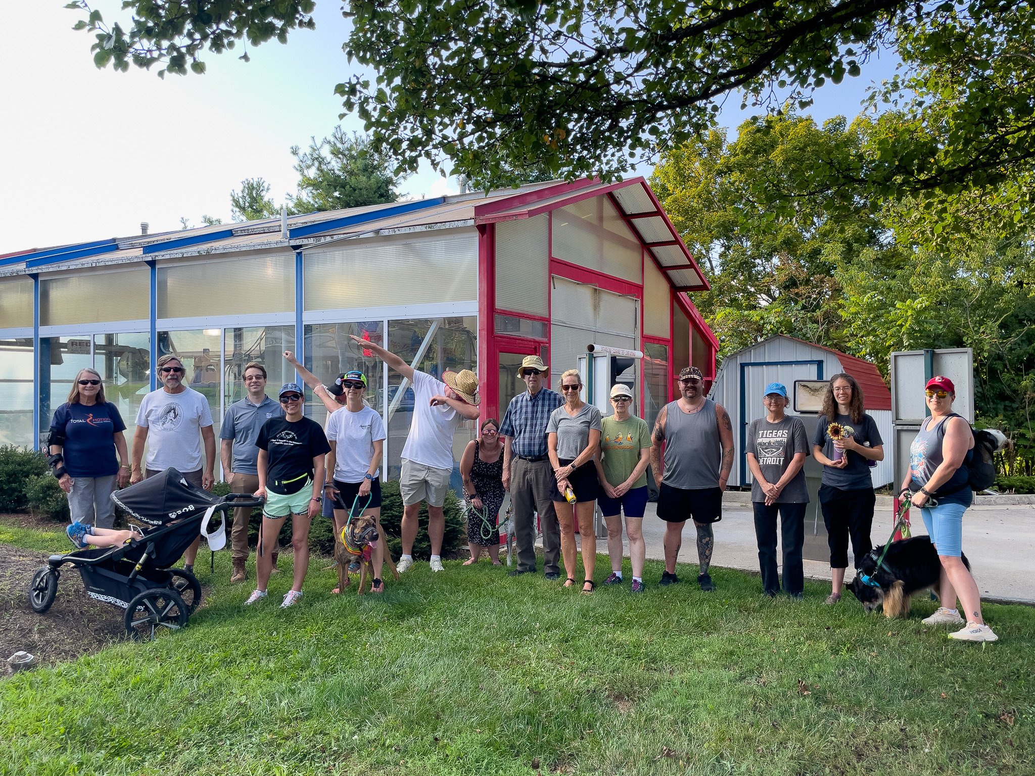 Group of people standing outdoors in front of a greenhouse, some holding leashes with dogs, others with sunflowers or backpacks, on a grassy area with trees in the background.