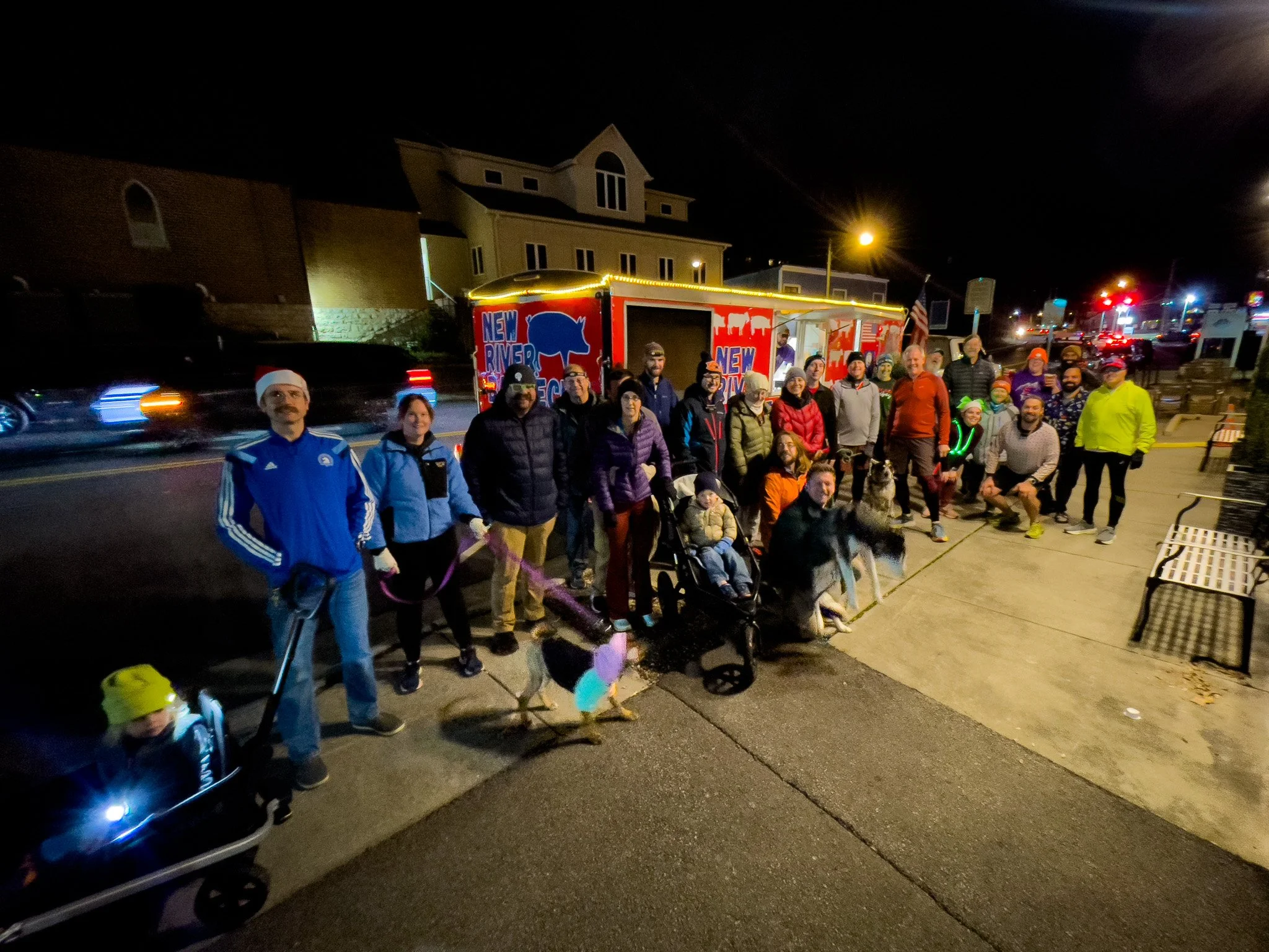 Group of people, some wearing holiday costumes, gathered on a sidewalk at night with dogs, a man in a Santa hat, and a decorated truck behind them.