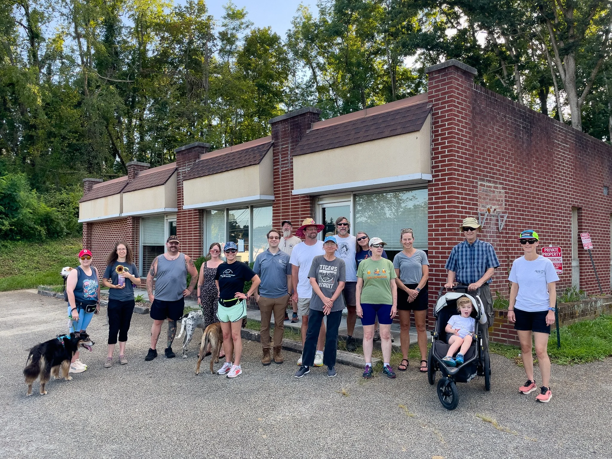 A group of people standing outside in front of a brick building with trees in the background. Some are holding dogs or standing near dogs, and a child is seated in a stroller.