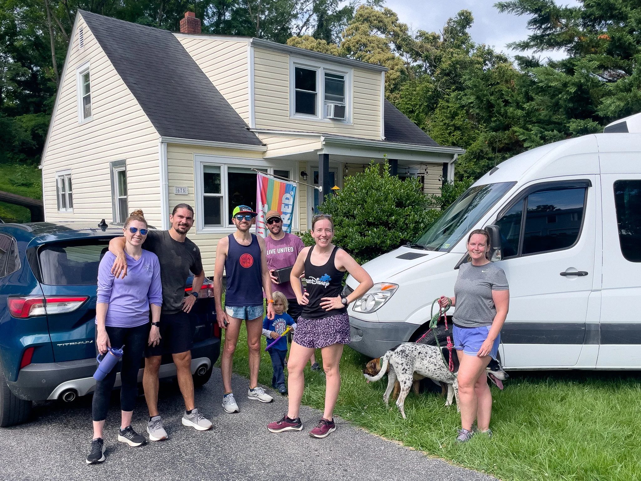 Group of seven people standing in front of a house with cars parked nearby, including a dog on a leash, smiling and posing for the photo on a sunny day.