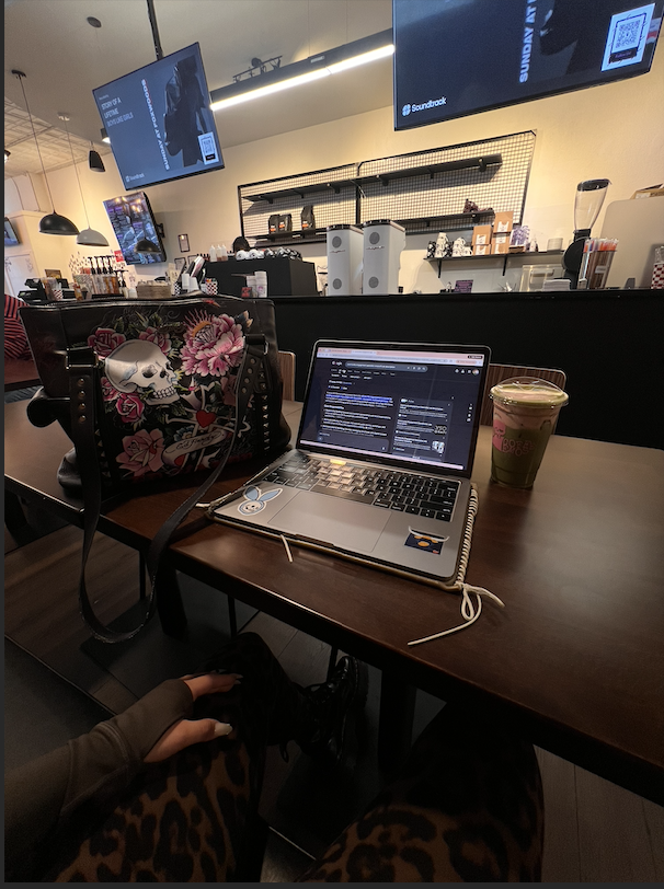 A table at a coffee shop with a laptop, a colorful bag with skull and floral designs, a cup of green drink, and a person's hand on their lap. The background features large monitors and coffee shop equipment.