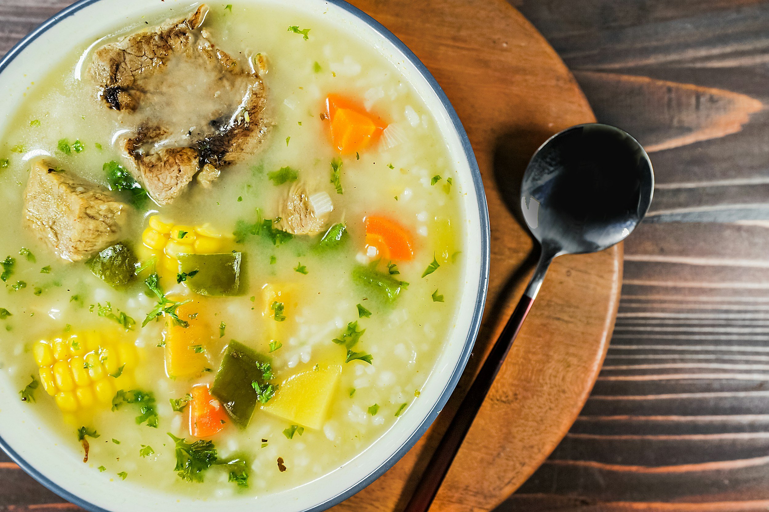 Bowl of hearty soup with meat, corn, carrots, green peppers, and rice on a wooden table.