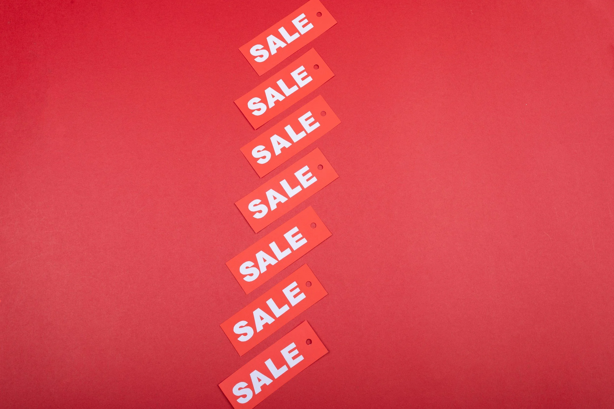 Seven sale tags with white letters are arranged diagonally on a red background for a vibrant sale display.