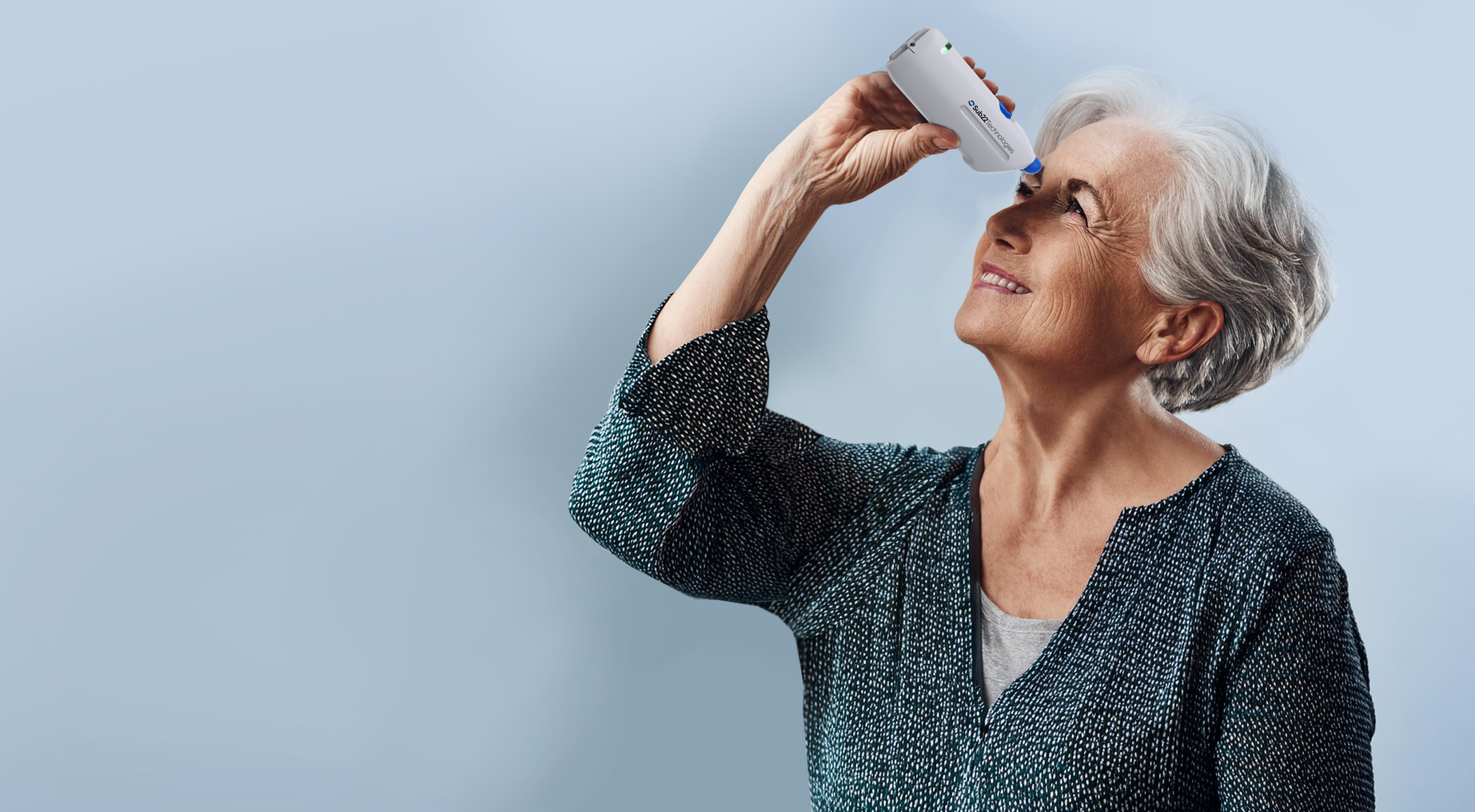 Older woman with gray hair smiling and looking up, using a medical device on her eye against a light blue background.