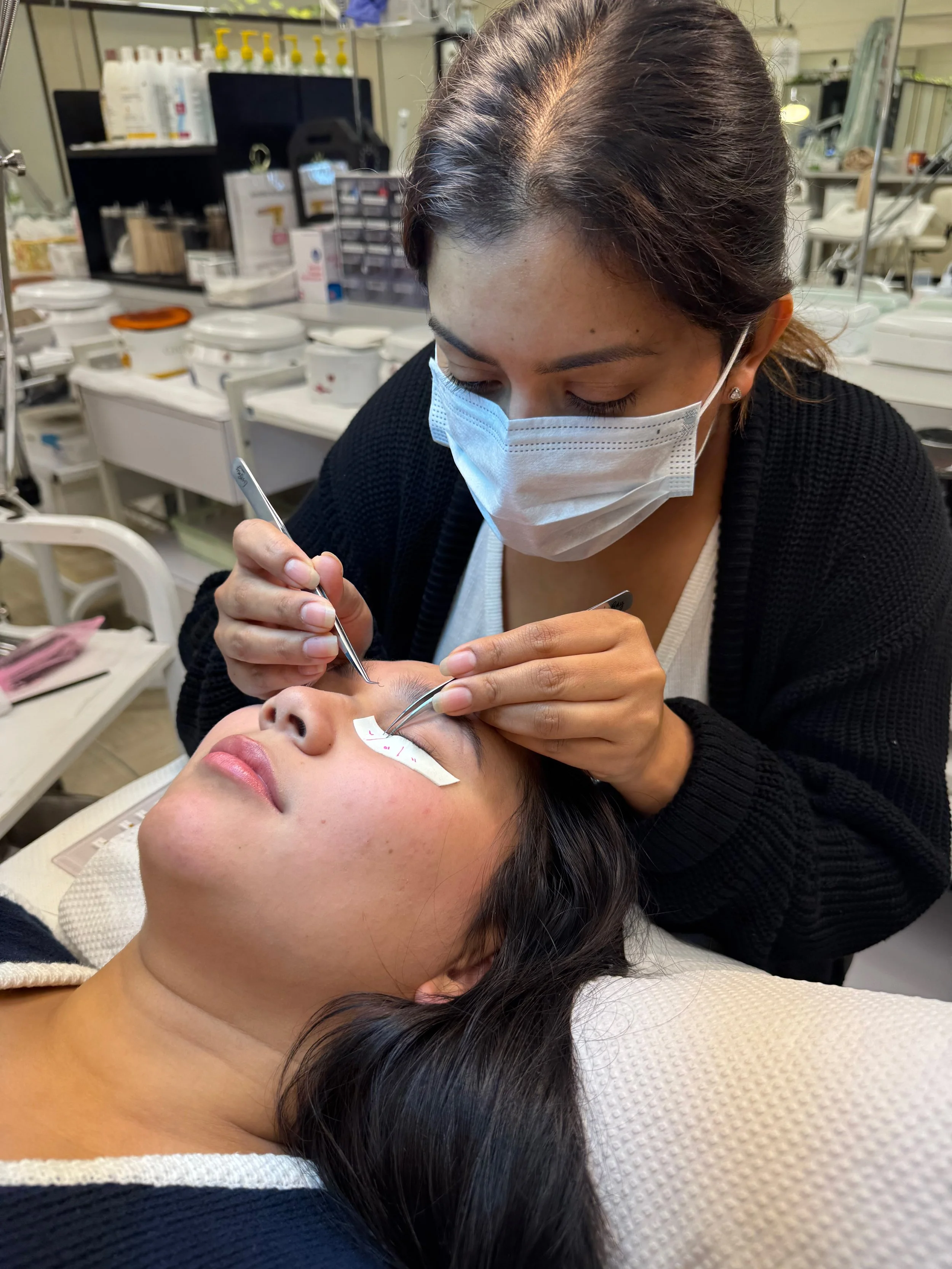 A woman receiving an eyelash extension or lash treatment from a technician in a clinic or beauty studio. The technician and client are wearing face masks.
