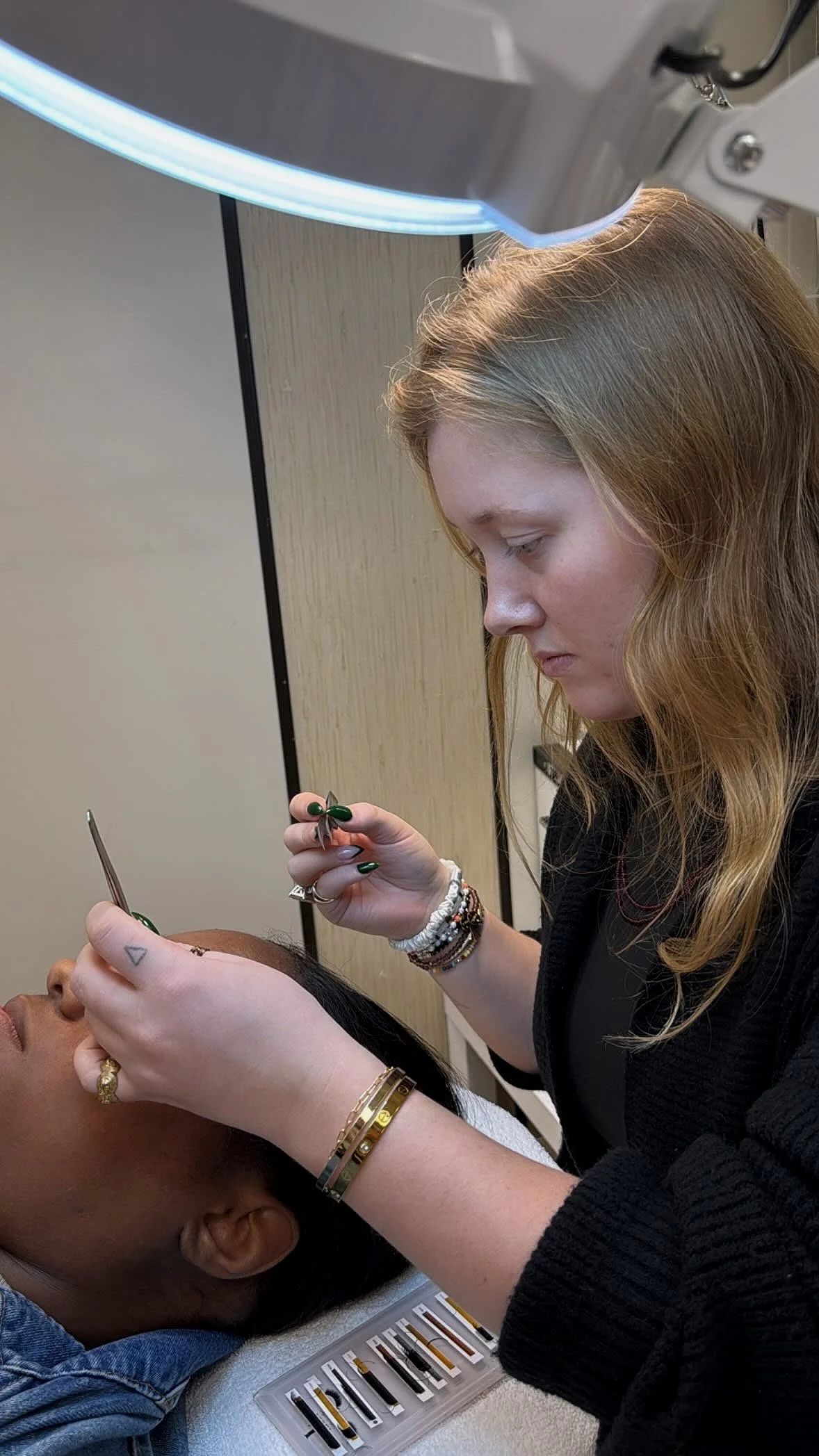 A woman provides a cosmetic or medical treatment to a woman lying down, using small tools near her eye under a bright overhead light.