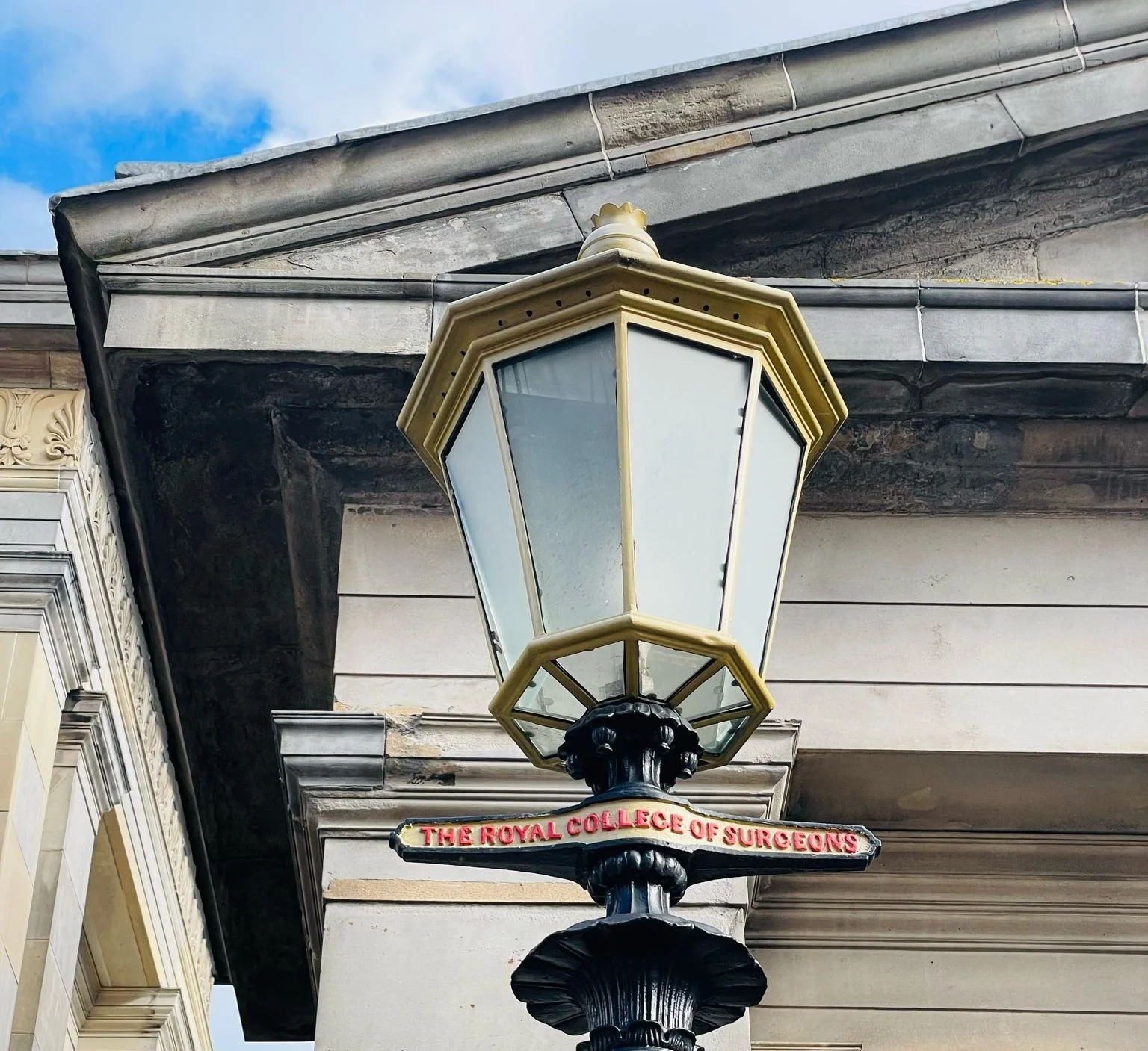 A vintage street lamp attached to a building with a sign reading 'The Royal College of Surgeons' beneath it, with ornate architecture and a cloudy sky in the background.