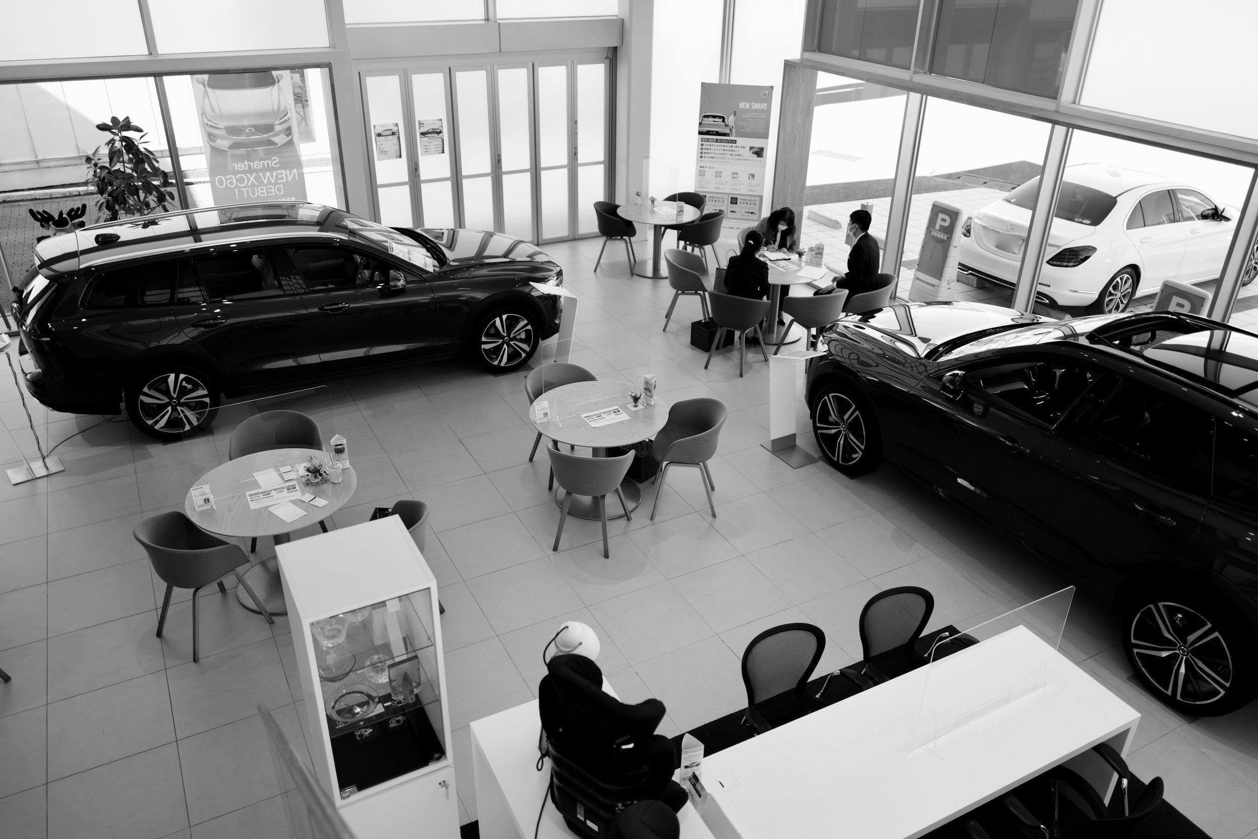 Black and white image of a car showroom with several vehicles on display and tables for customer consultations.