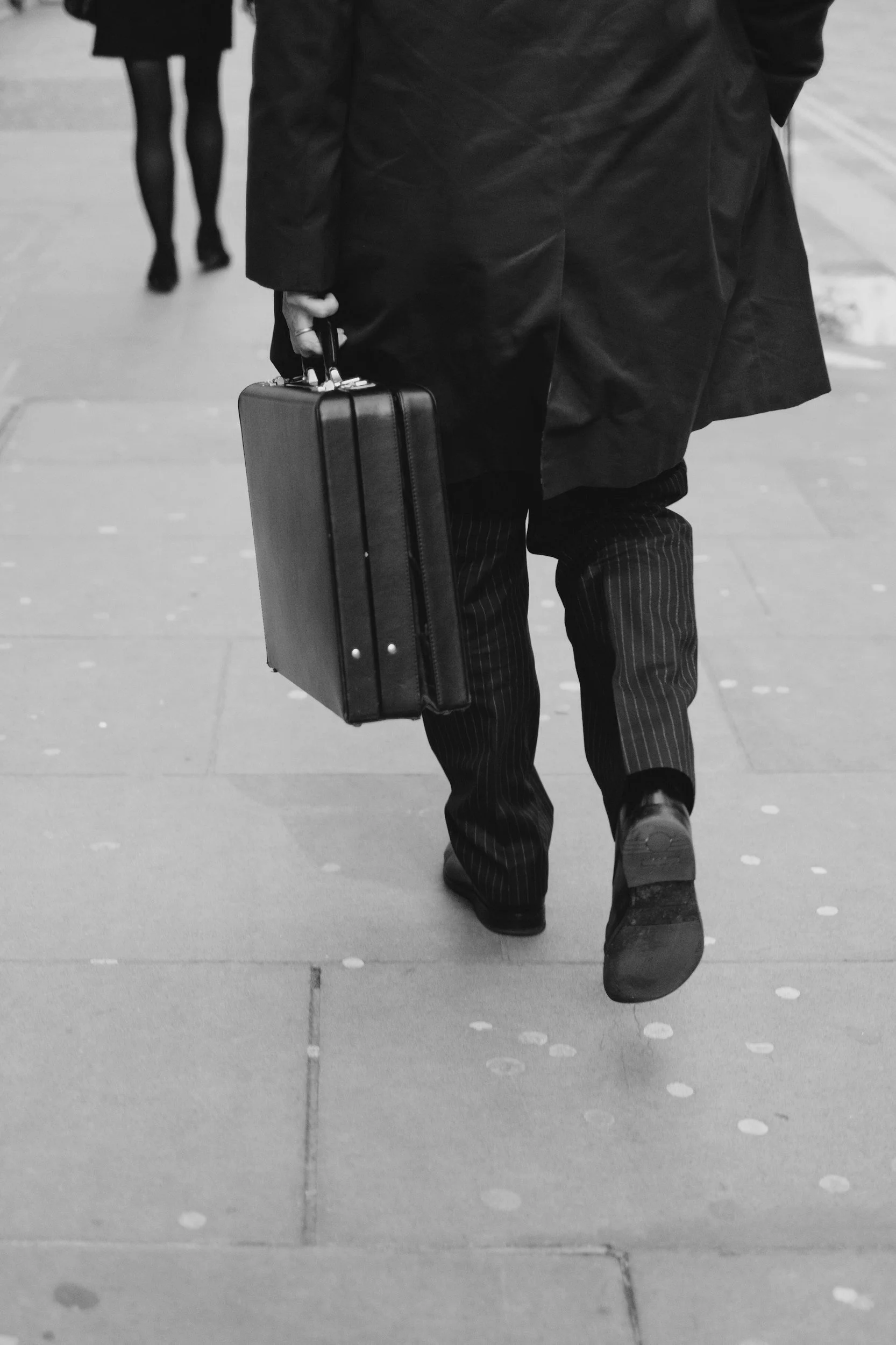 Black and white photo of a man in a suit walking on a pavement carrying a briefcase.