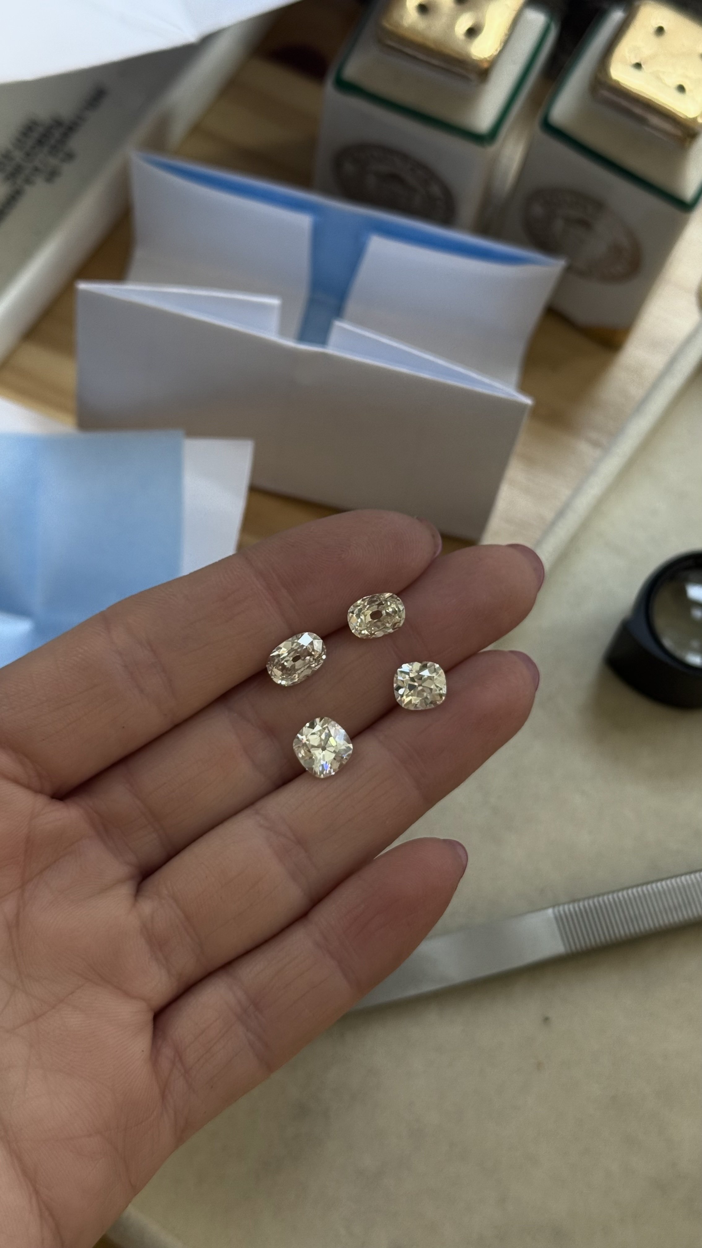 Hand holding four large, oval-shaped, clear gemstones or diamonds, with jewelry supplies and boxes in the background.