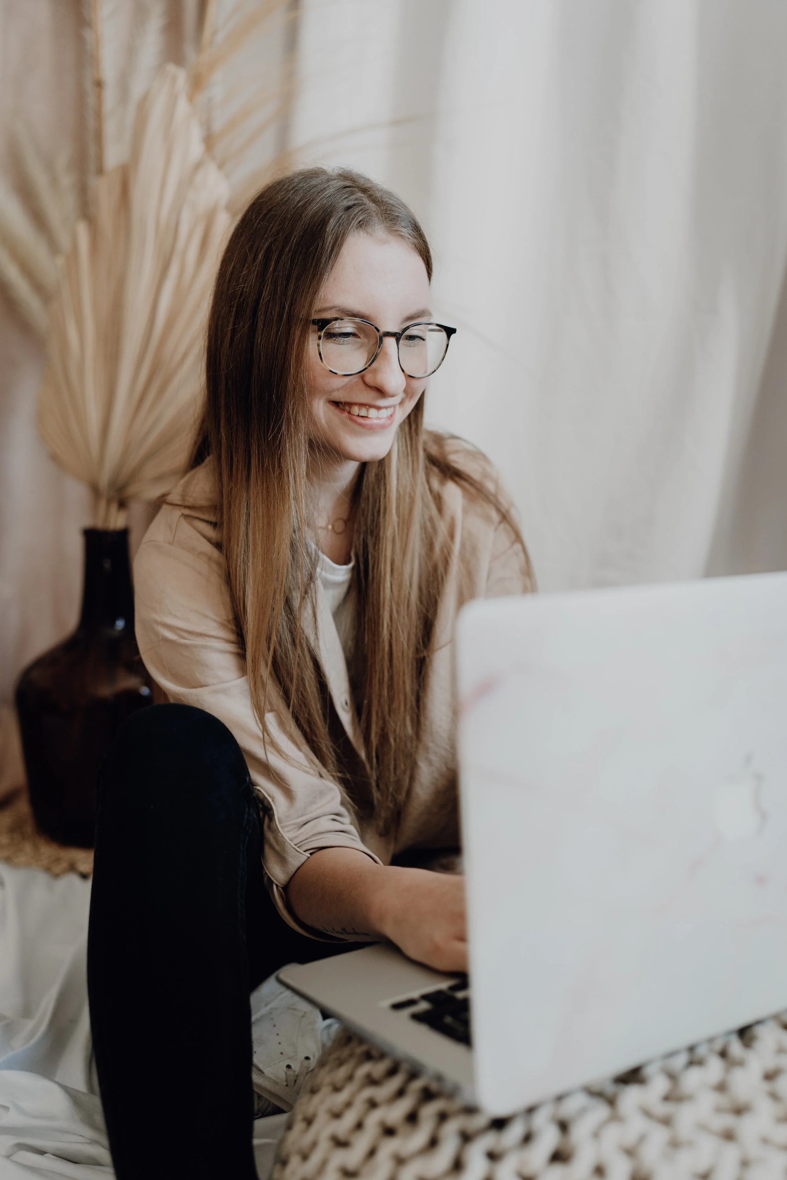 Junge Frau mit Brille arbeitet lächelnd auf einem Laptop in einem gemütlichen Raum mit getrockneten Pflanzen im Hintergrund.