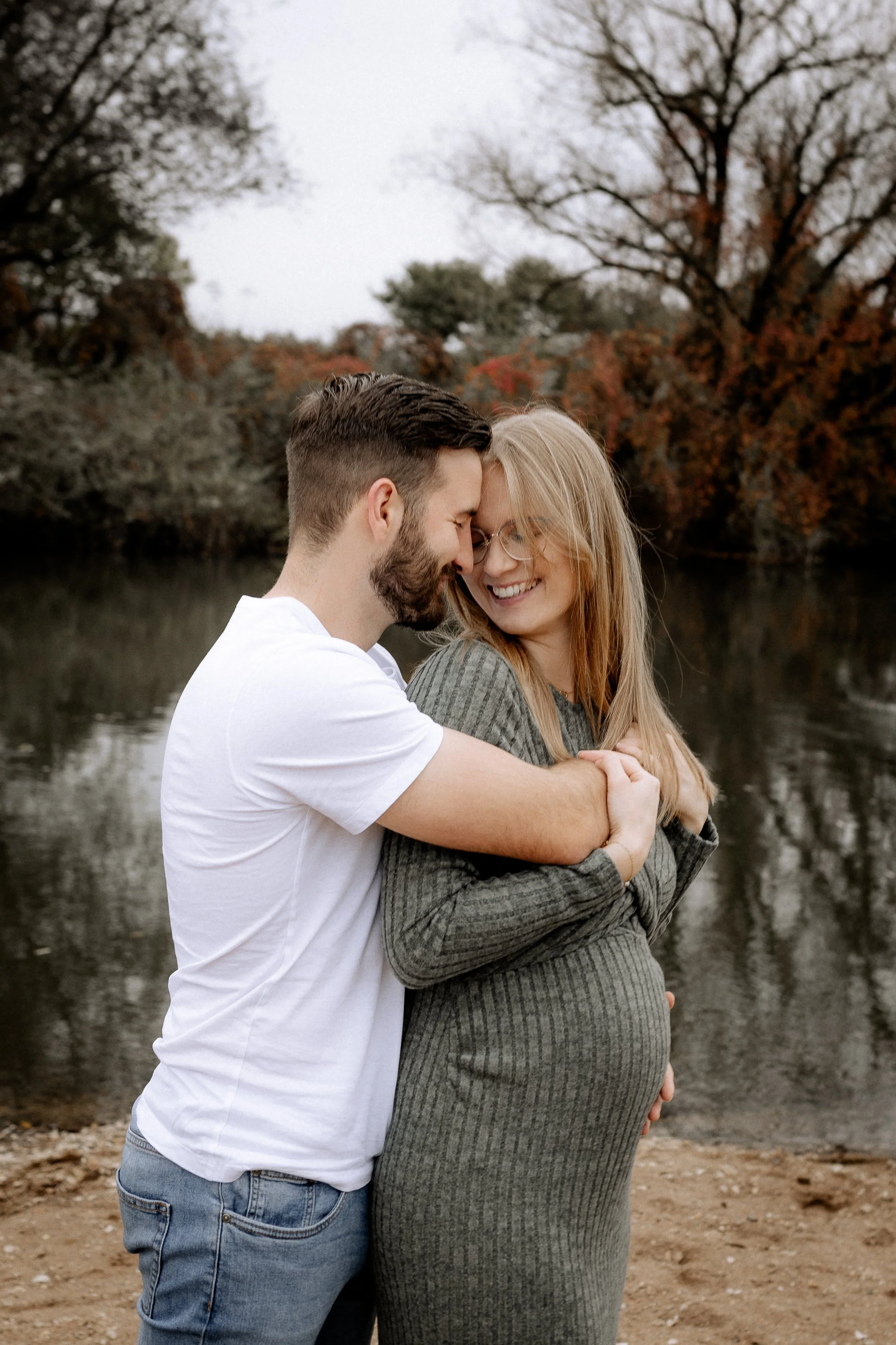 Ein glückliches Paar umarmt sich am Fluss im Herbst, die schwangere Frau lacht, während sie den Kopf an den Mann lehnt.