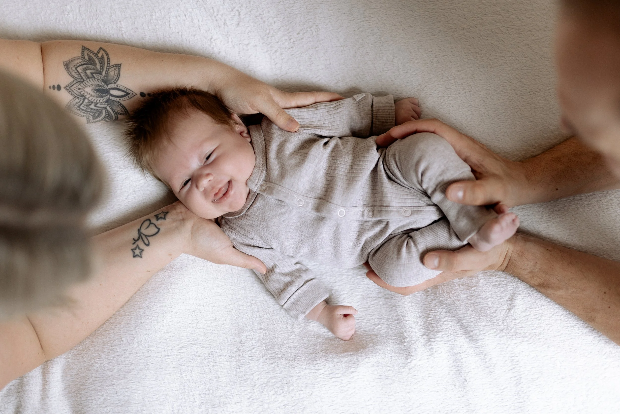 Ein glückliches Baby liegt auf einem weißen Tuch, von zwei Erwachsenen liebevoll gehalten.
