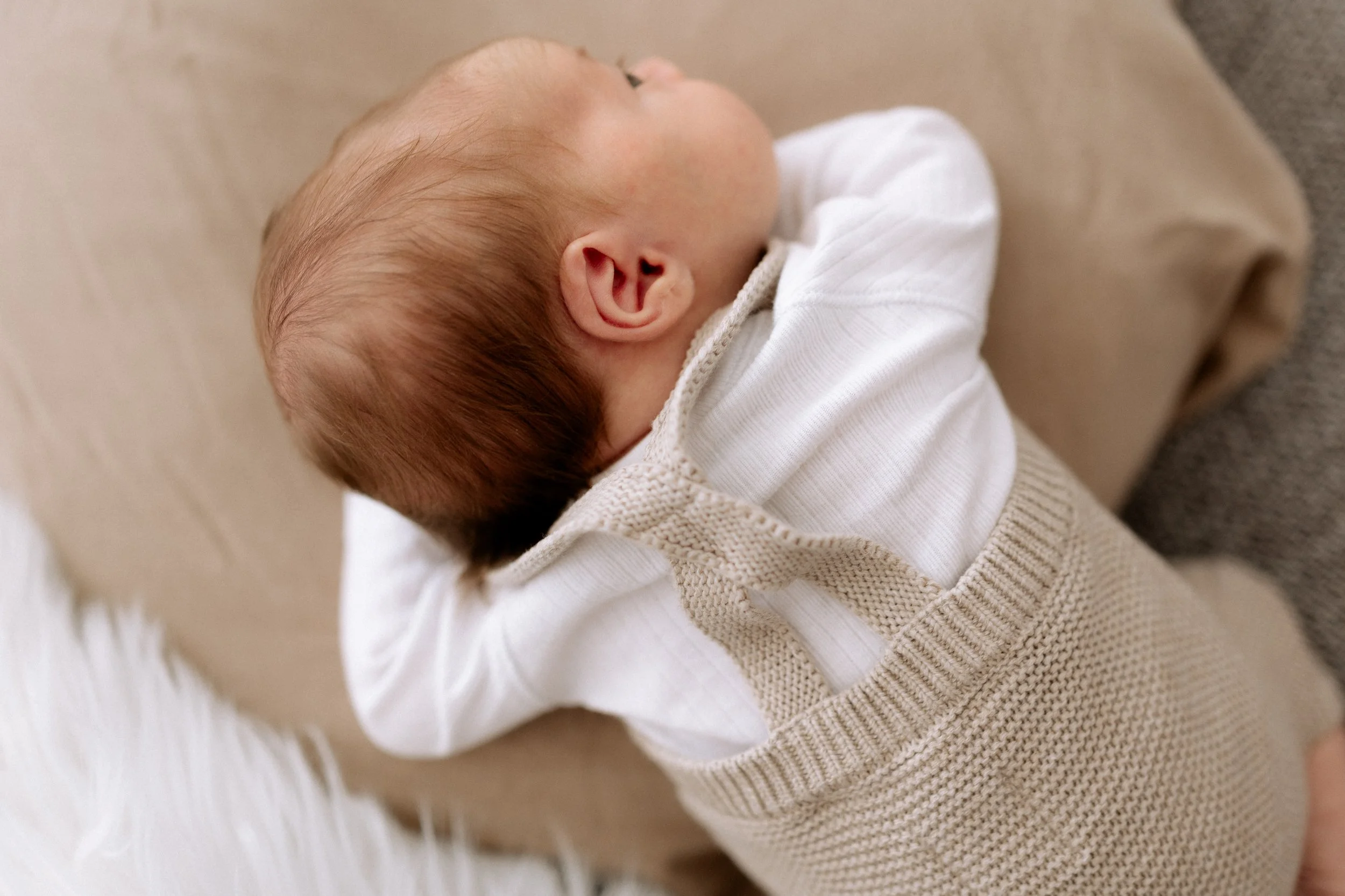 Ein schlafender Baby liegt auf einer beige Decke, trägt eine weiße Kleidung und einen beigefarbenen gestrickten Pullover