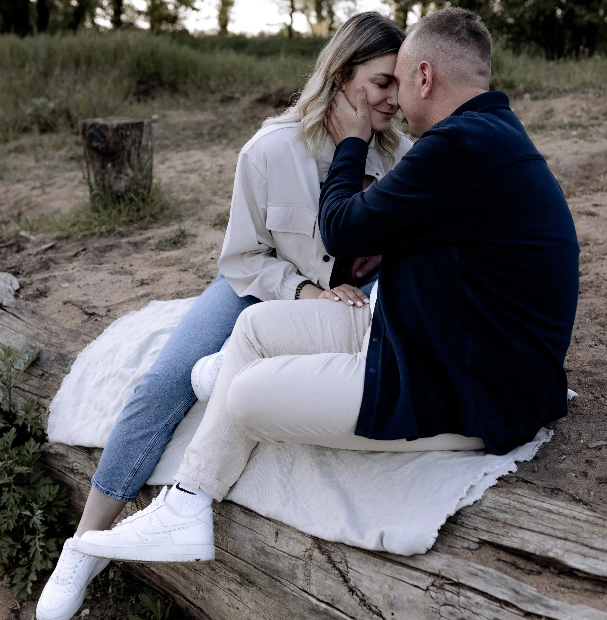 Ein Mann und eine Frau sitzen eng beieinander auf einer Decke im Freien, berühren sich liebevoll und lächeln.