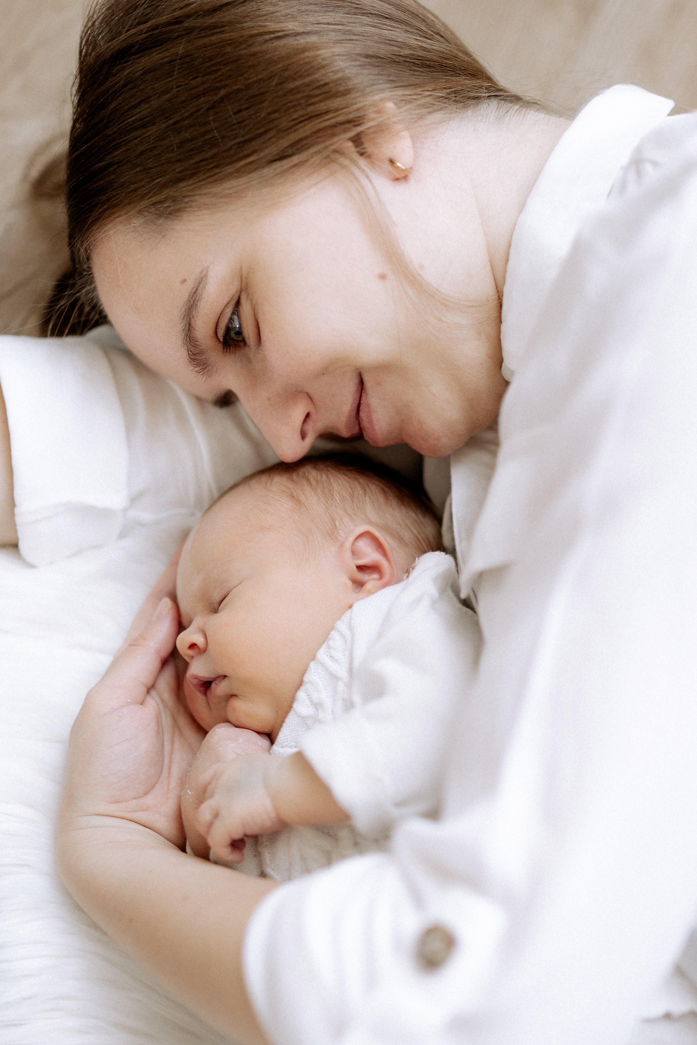 Eine Mutter liegt auf dem Bett und hält ihr schlafendes neugeborenes Baby liebevoll im Arm, beide tragen weiße Kleidung.