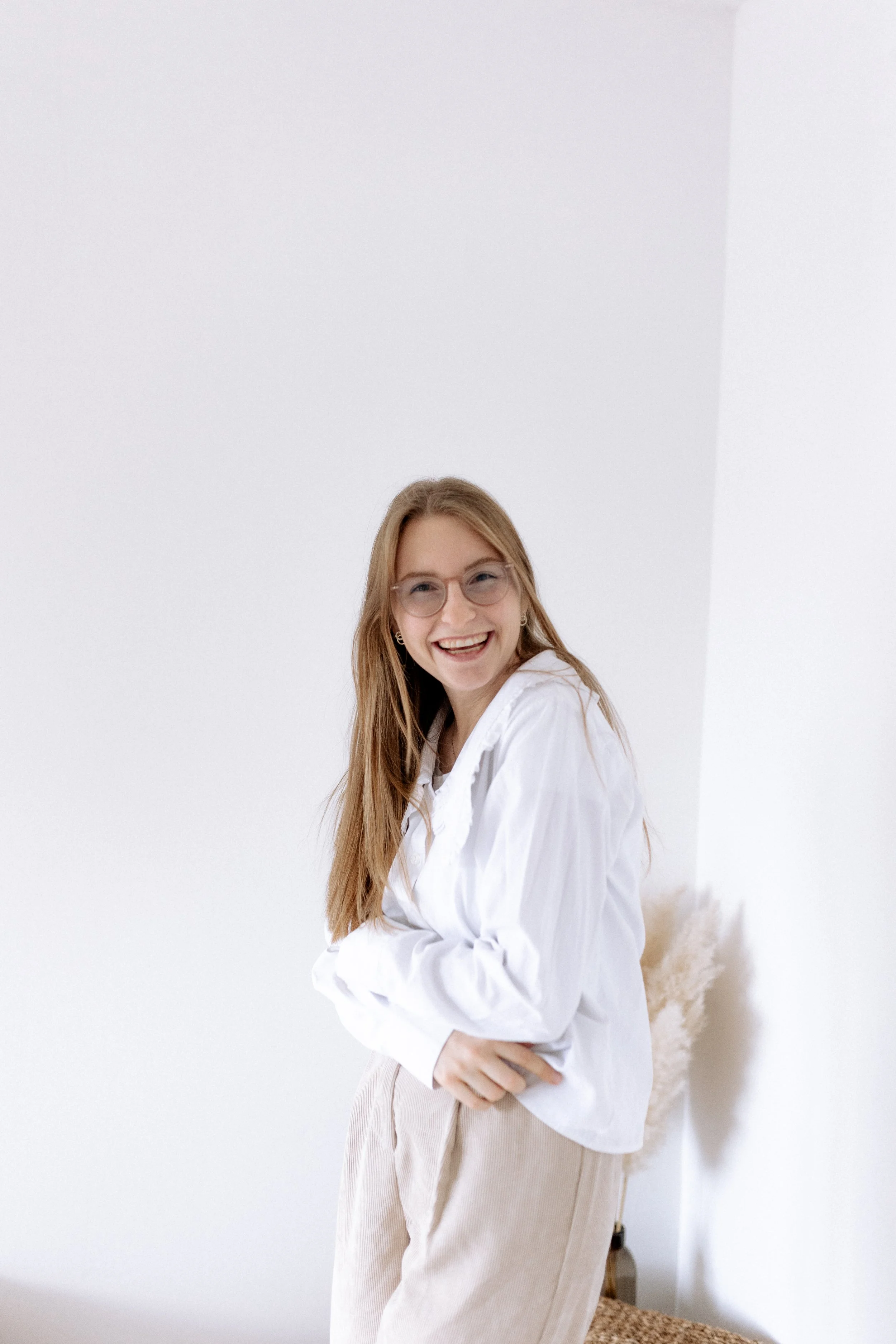 Lächelnde junge Frau mit Brille, hellbraunes Haar, in weißer Jacke und bequemen beigen Hosen, stehend vor weißer Wand in einem modernen Raum.