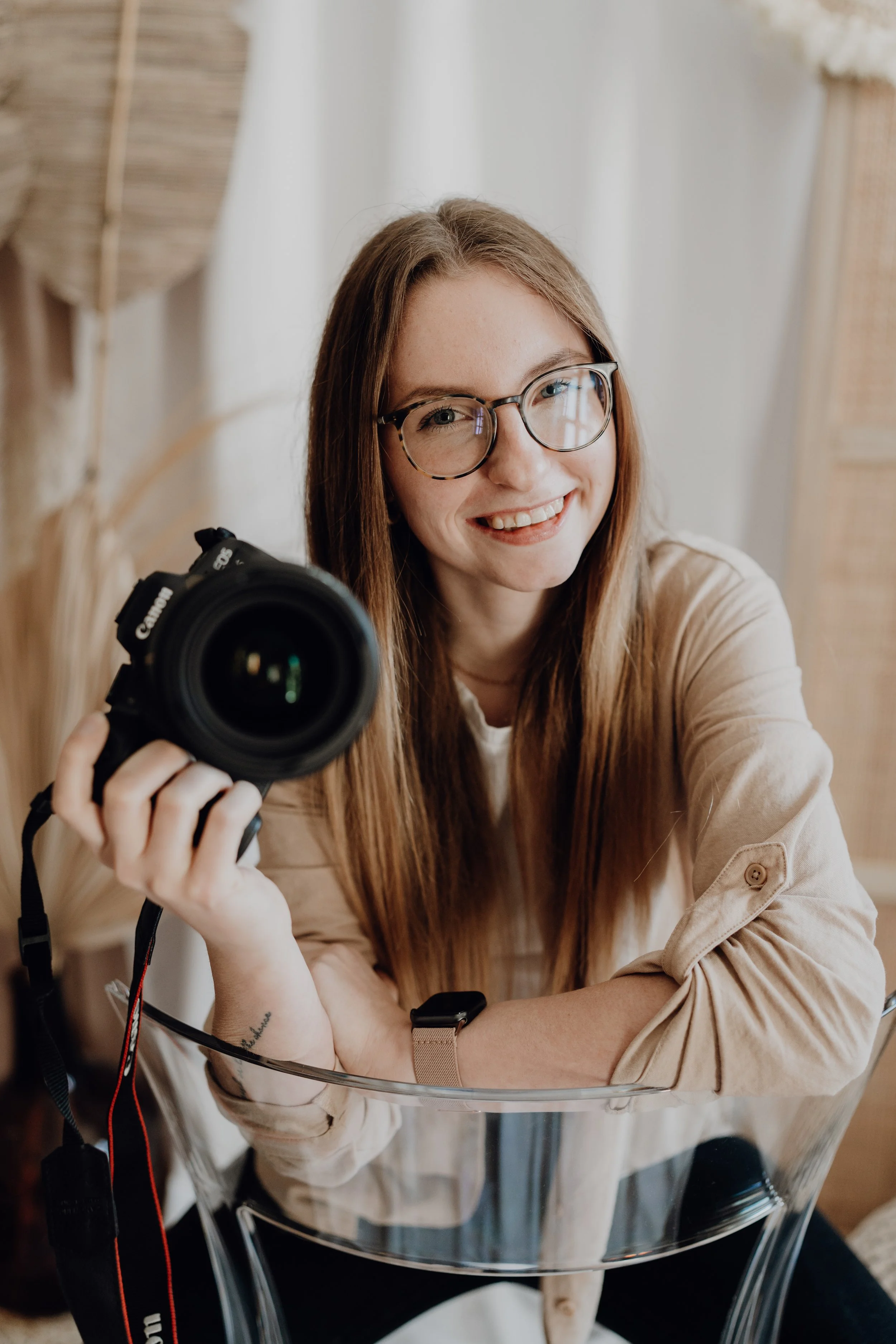 Junge Frau mit Brille hält eine DSLR-Kamera, sitzt auf einem transparenten Stuhl, lächelt, hat lange braune Haare, trägt ein beiges Jacke und befindet sich in einem hellen Raum mit natürlichem Licht.