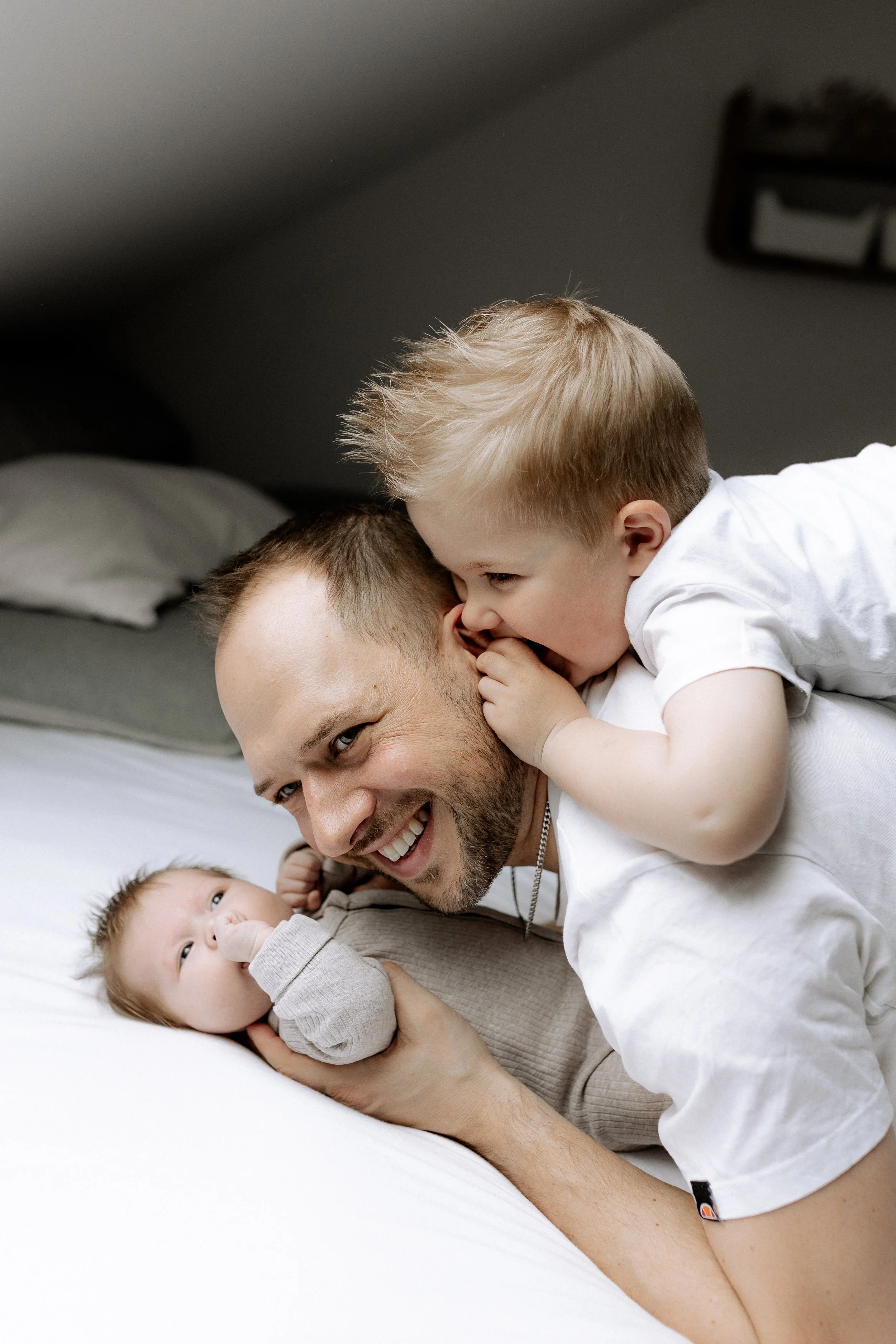 Ein Vater mit seinen zwei kleinen Kindern, eines auf dem Bauch und das andere auf seiner Schulter, im Schlafzimmer.