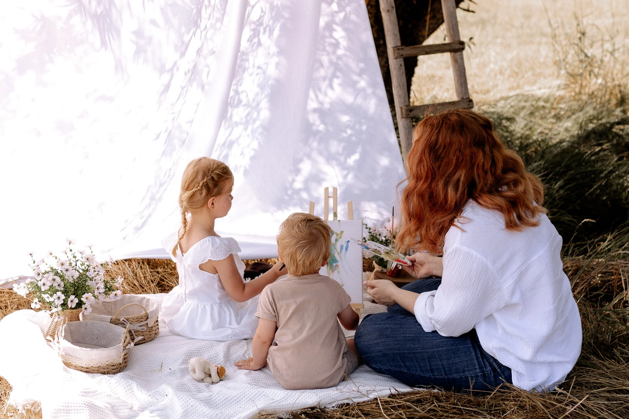 Eine Frau und zwei Kinder malen gemeinsam im Freien vor einer weißen Leinwand, mit Büchern, Blumen und einem Teddy auf einer Decke, bei Sonnenlicht.