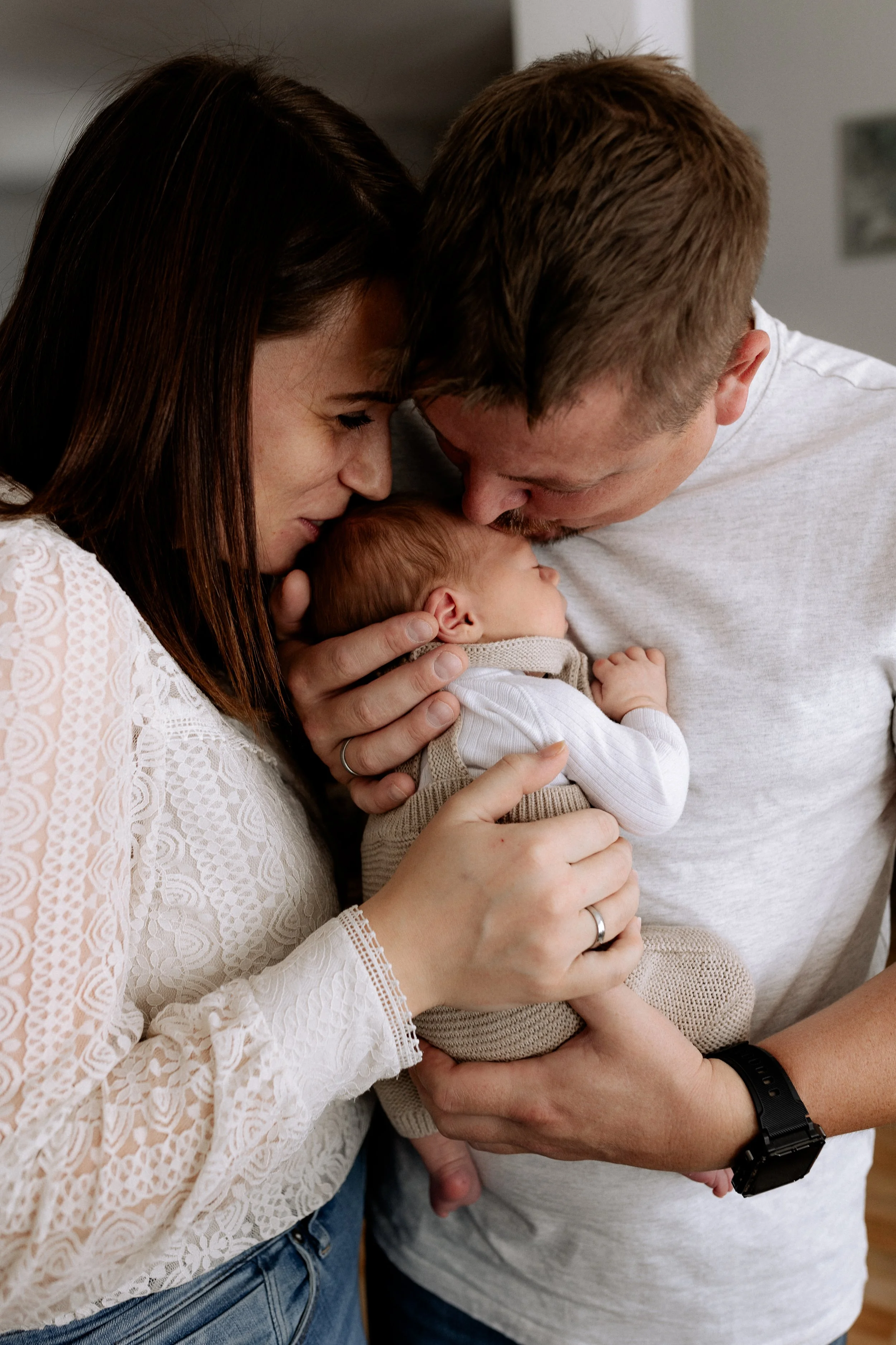 Ein Paar hält ein schlafendes Baby in den Armen, beide berühren ihre Stirn und schauen liebevoll auf das Kind, in einem gemütlichen Zuhause.