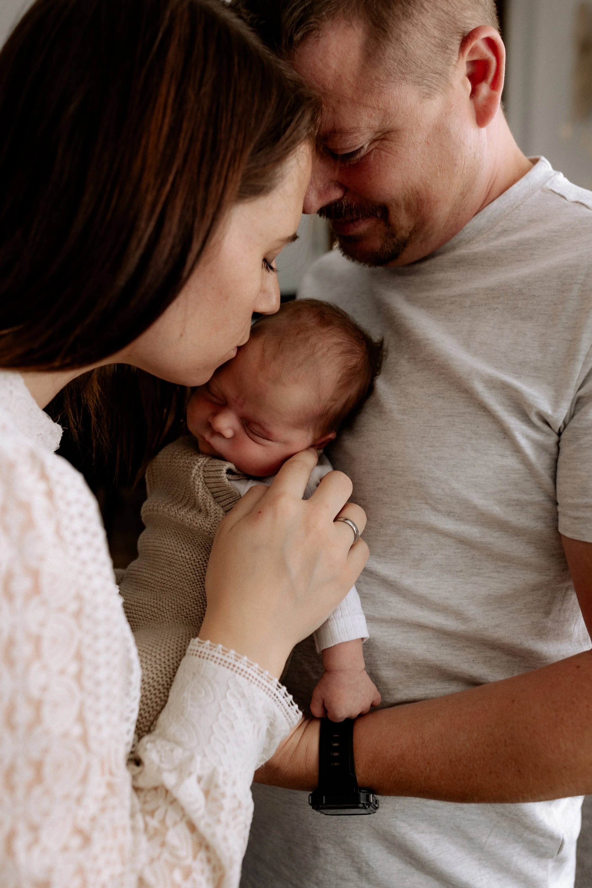 Ein Baby wird von einer Frau sanft gehalten, während ein Mann lächelnd dabei ist, alle drei berühren sich mit ihren Stirnen in einer liebevollen Geste.