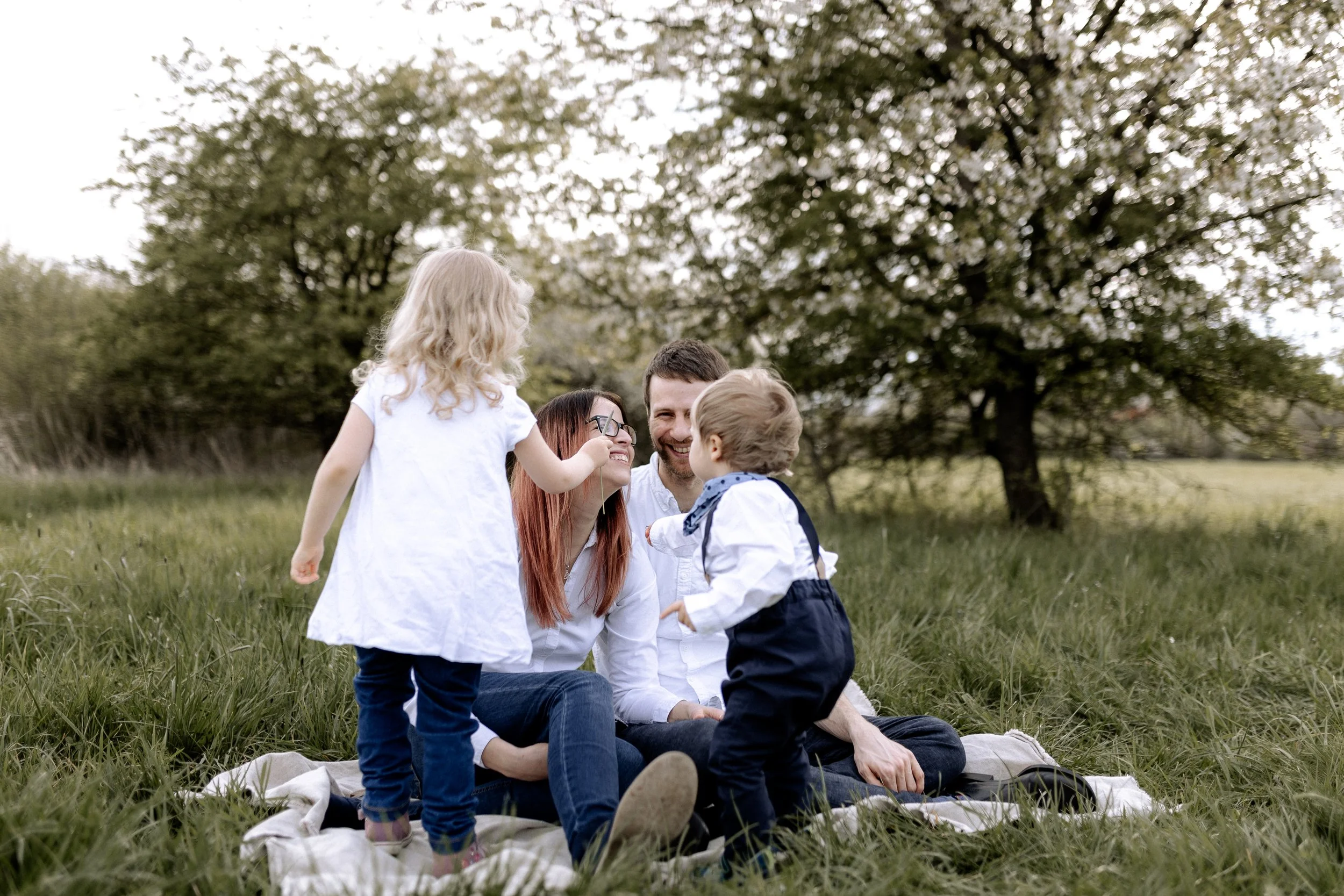 Familie mit zwei kleinen Kindern beim Picknick im Grünen, sitzend auf einer Decke, lachend vor einem Baum