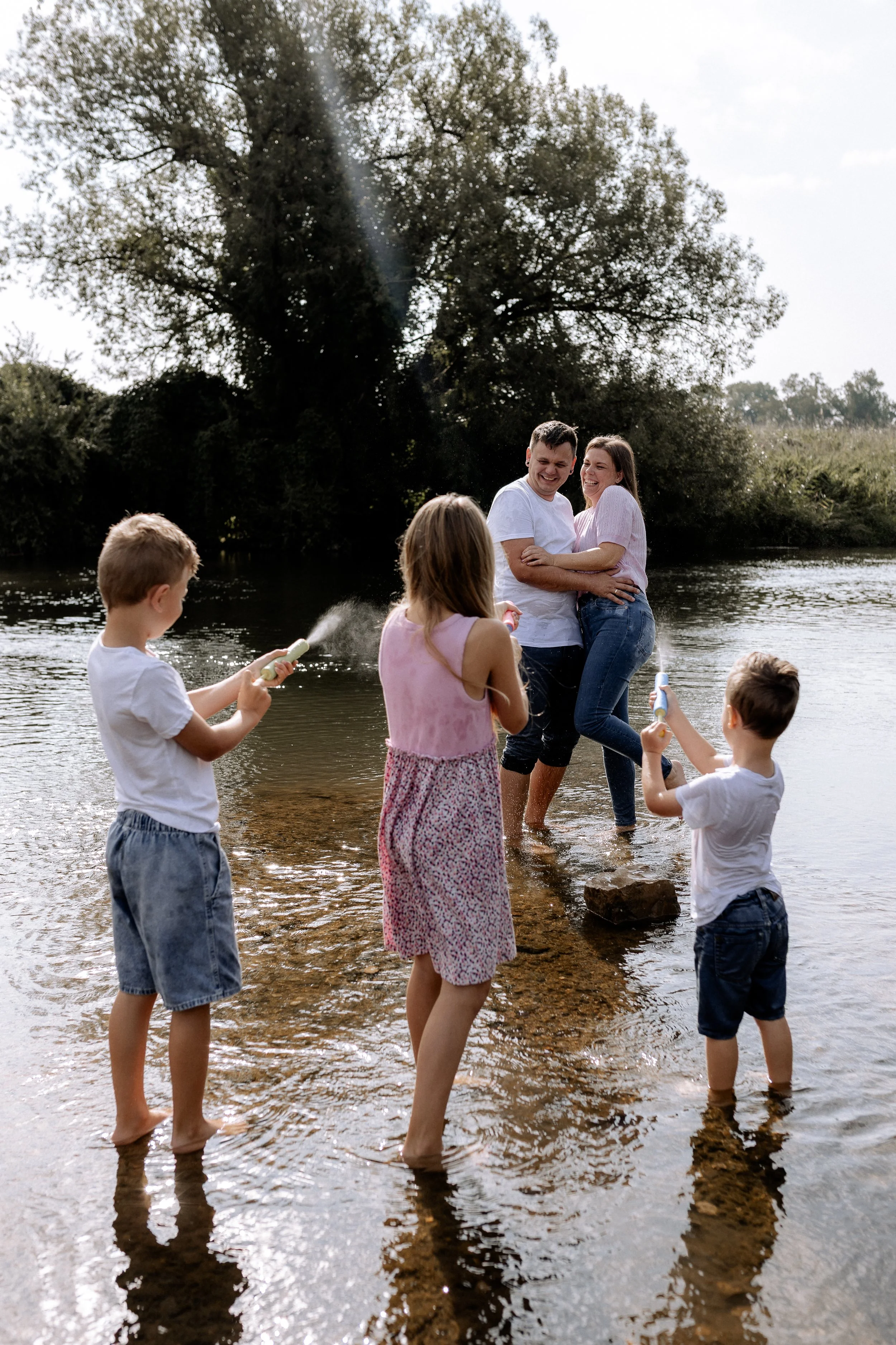 Familie spielt fröhlich im Wasser eines Flusses, während die Eltern die Kinder umarmen, bei sonnigem Wetter in der Natur.