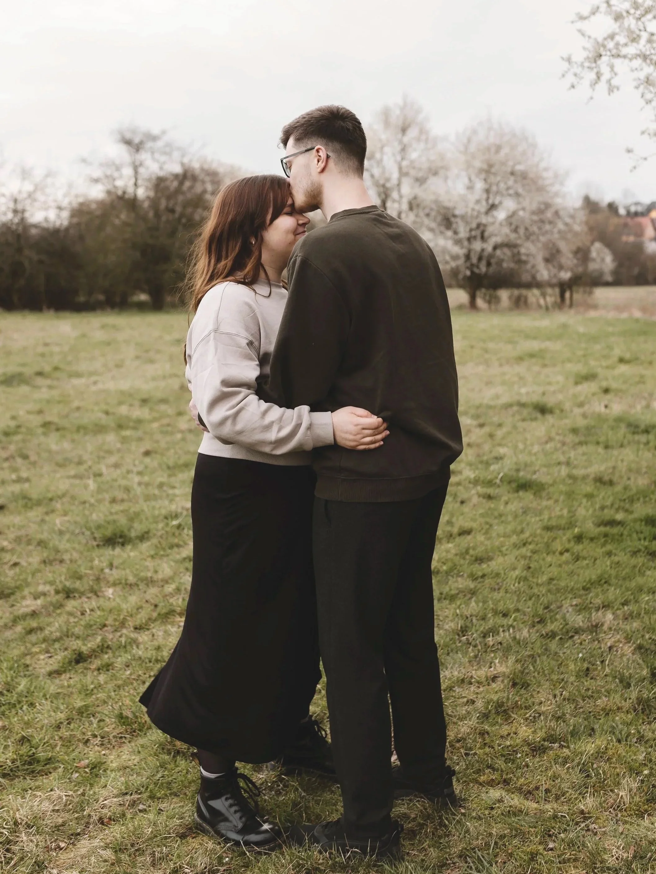 Ein glückliches Paar umarmt sich auf einer grünen Wiese im Freien, mit blühenden Bäumen im Hintergrund, an einem bewölkten Tag.