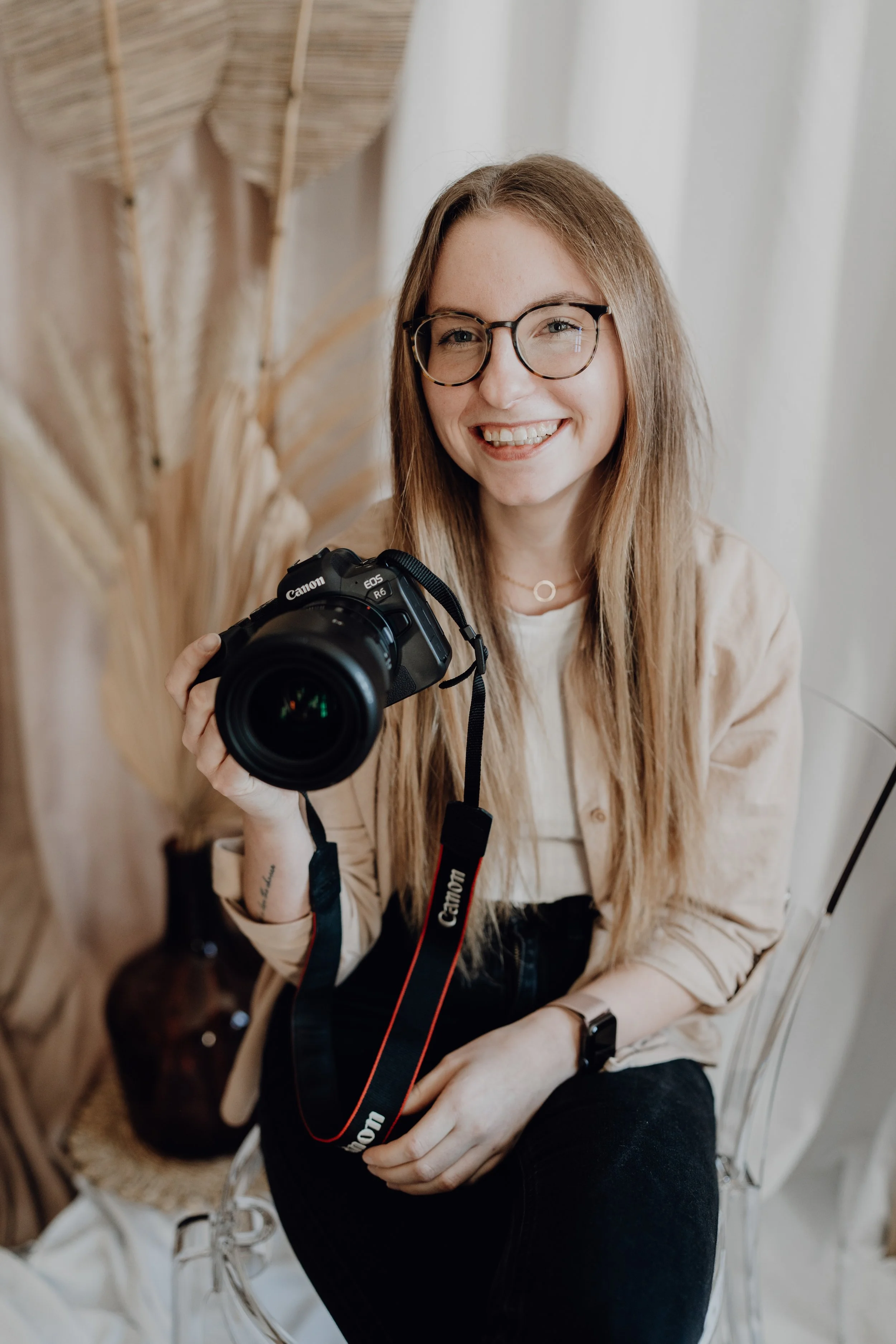 Eine lachende junge Frau mit Brille hält eine Kamera in der Hand und sitzt auf einem transparenten Stuhl, im Hintergrund sind getrocknete Pflanzen und ein Vase sichtbar.