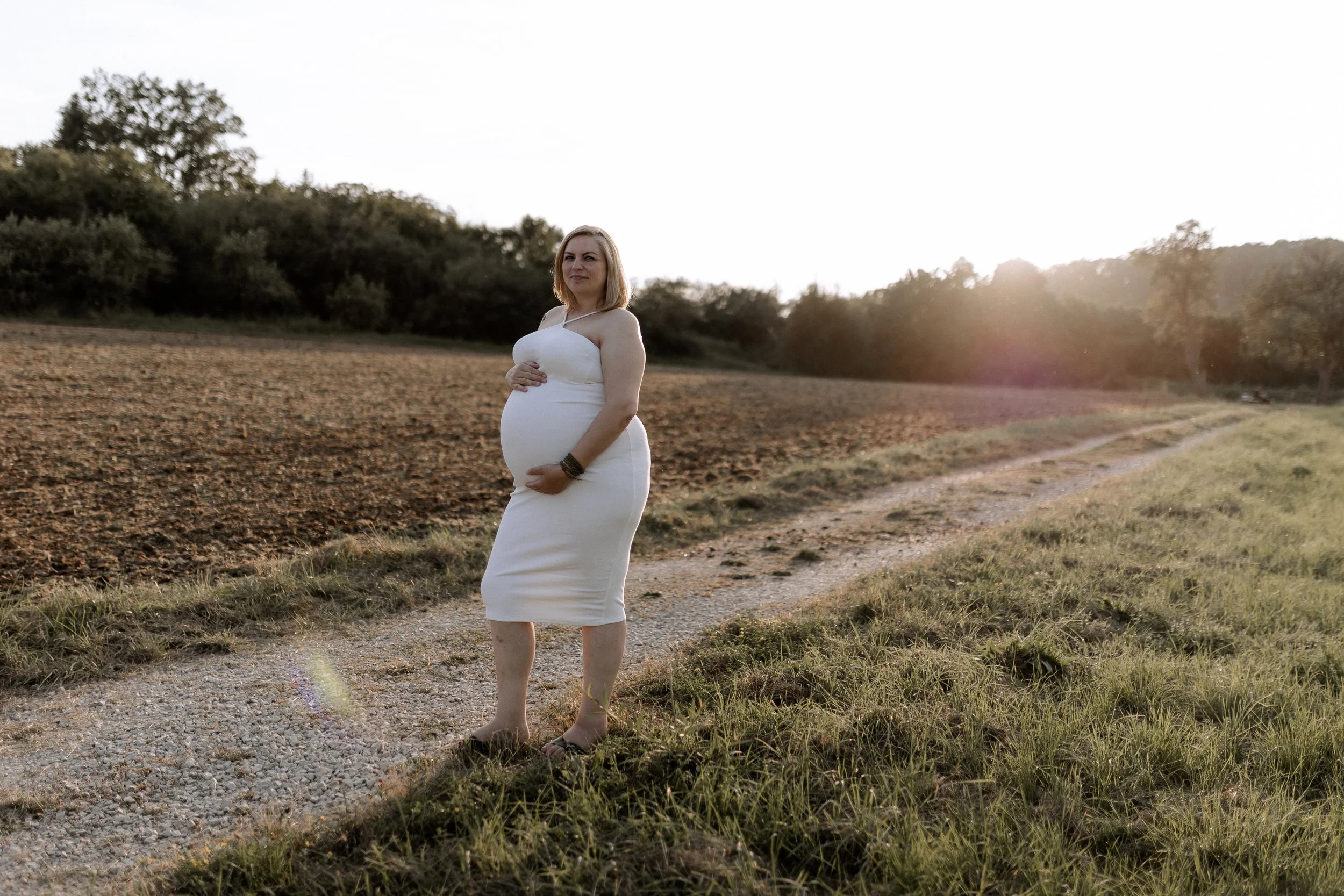 Schwangere Frau in weißem Kleid steht auf einem Feldweg bei Sonnenuntergang, umgeben von Natur.