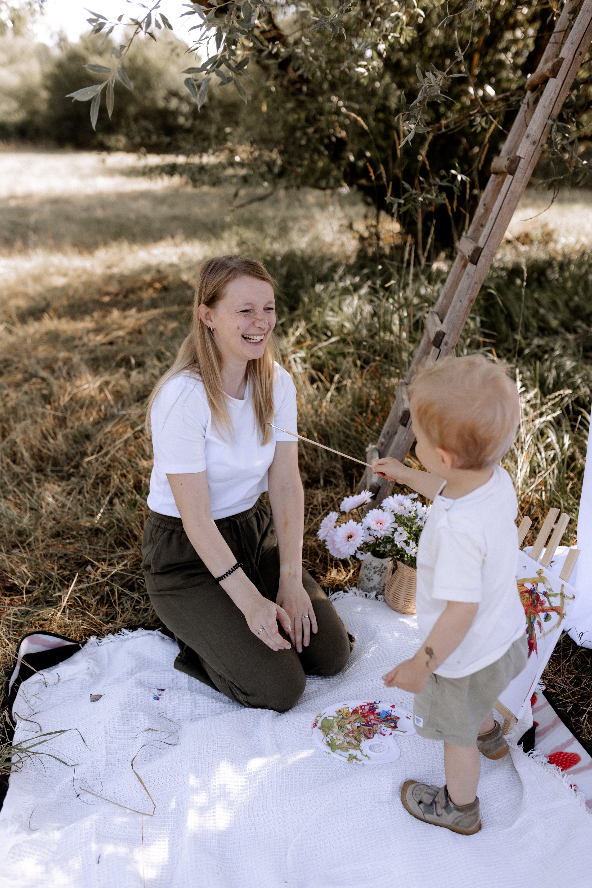 Eine lachende Frau und ein kleines Kind bei einem Picknick im Freien, mit Blumen, einem Korb und einer Leiter im Hintergrund.