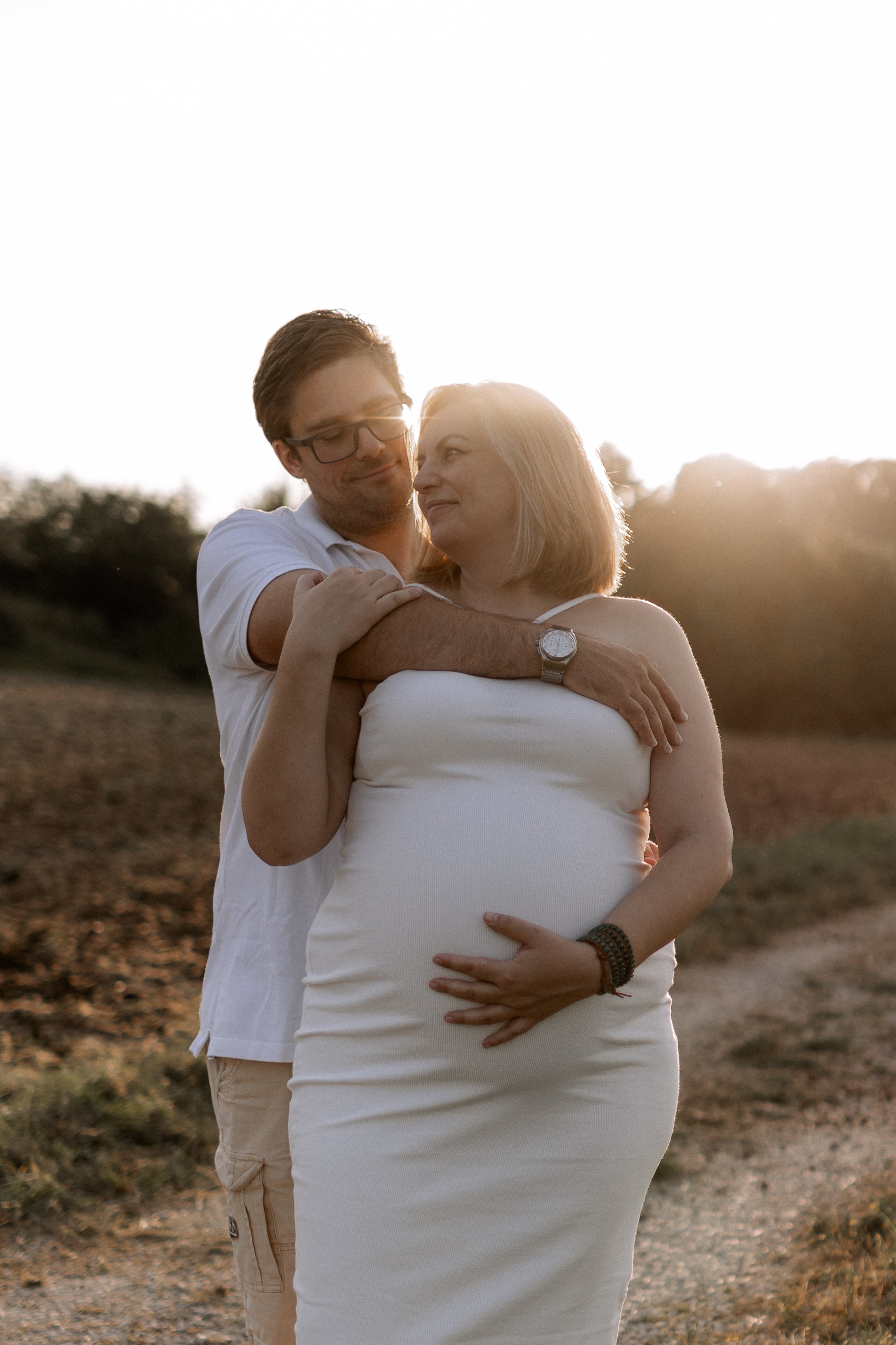 Ein Paar in einem Feld bei Sonnenuntergang, die Frau ist schwanger und wird von ihrem Partner liebevoll umarmt.