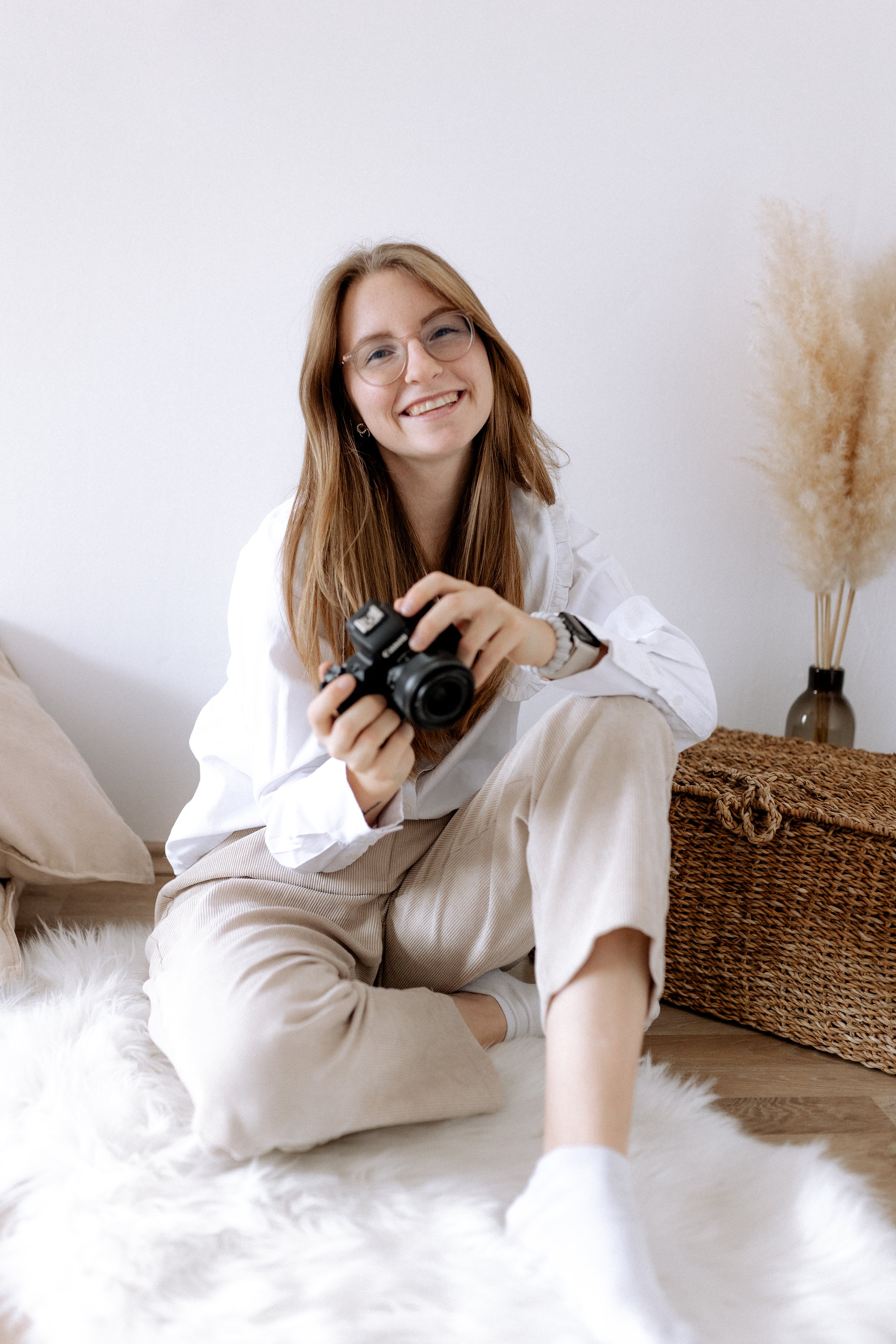 Junge Frau mit Brille sitzt auf Teppich und hält eine Kamera, lächelnd in einem Zimmer mit weißer Wand und Dekoration.