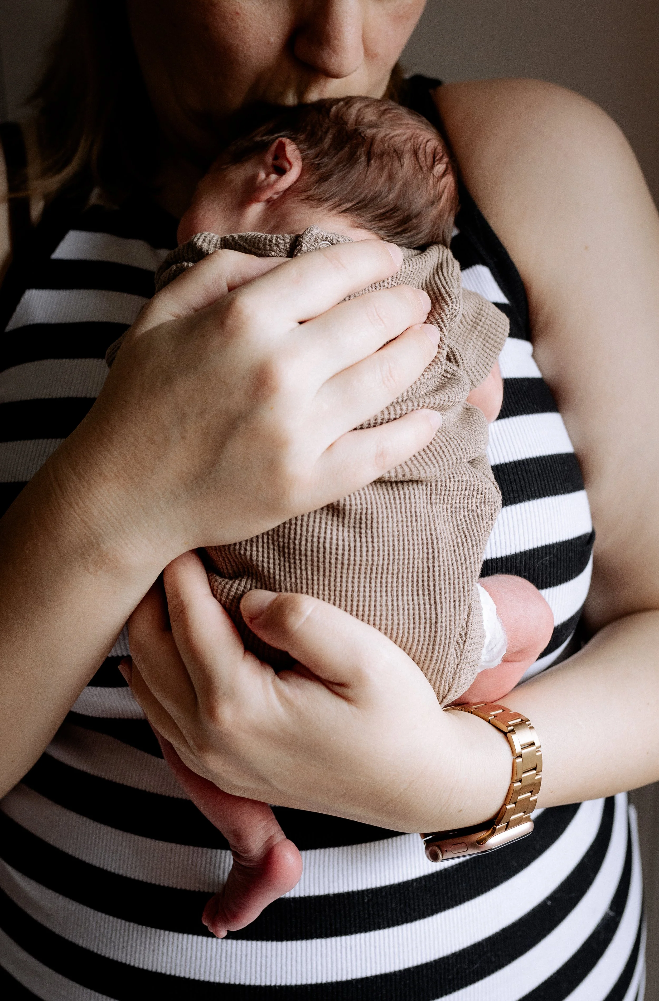 Eine Frau hält ein neugeborenes Baby eng an ihrer Brust, trägt ein schwarzes und weiß gestreiftes Top, trägt einen goldfarbenen Armband und schaut nach unten auf das Baby, das in einem braunen Kleid gekleidet ist.