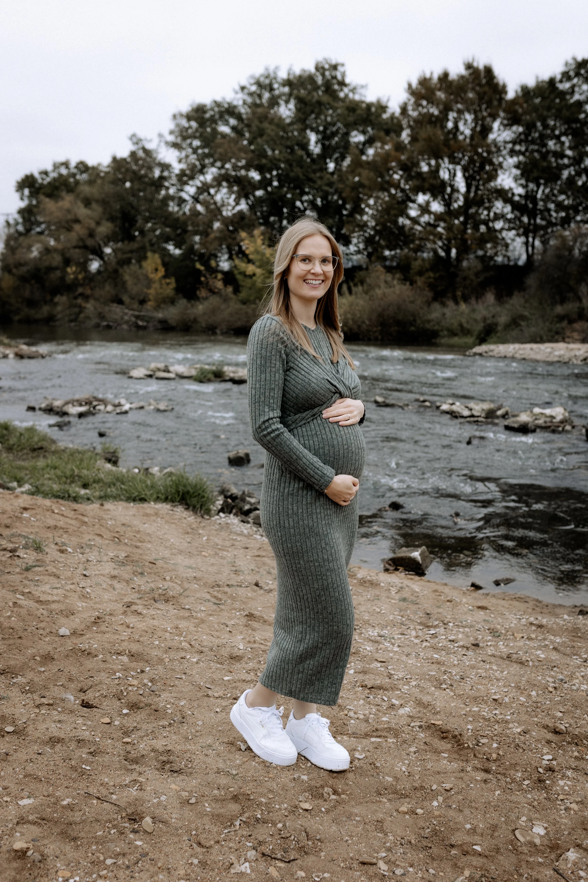 Schwangere Frau steht am Flussufer, trägt grauen langärmeliges Kleid und weiße Turnschuhe, lächelt in die Kamera, im Hintergrund Bäume und Wasser, bewölkter Himmel.