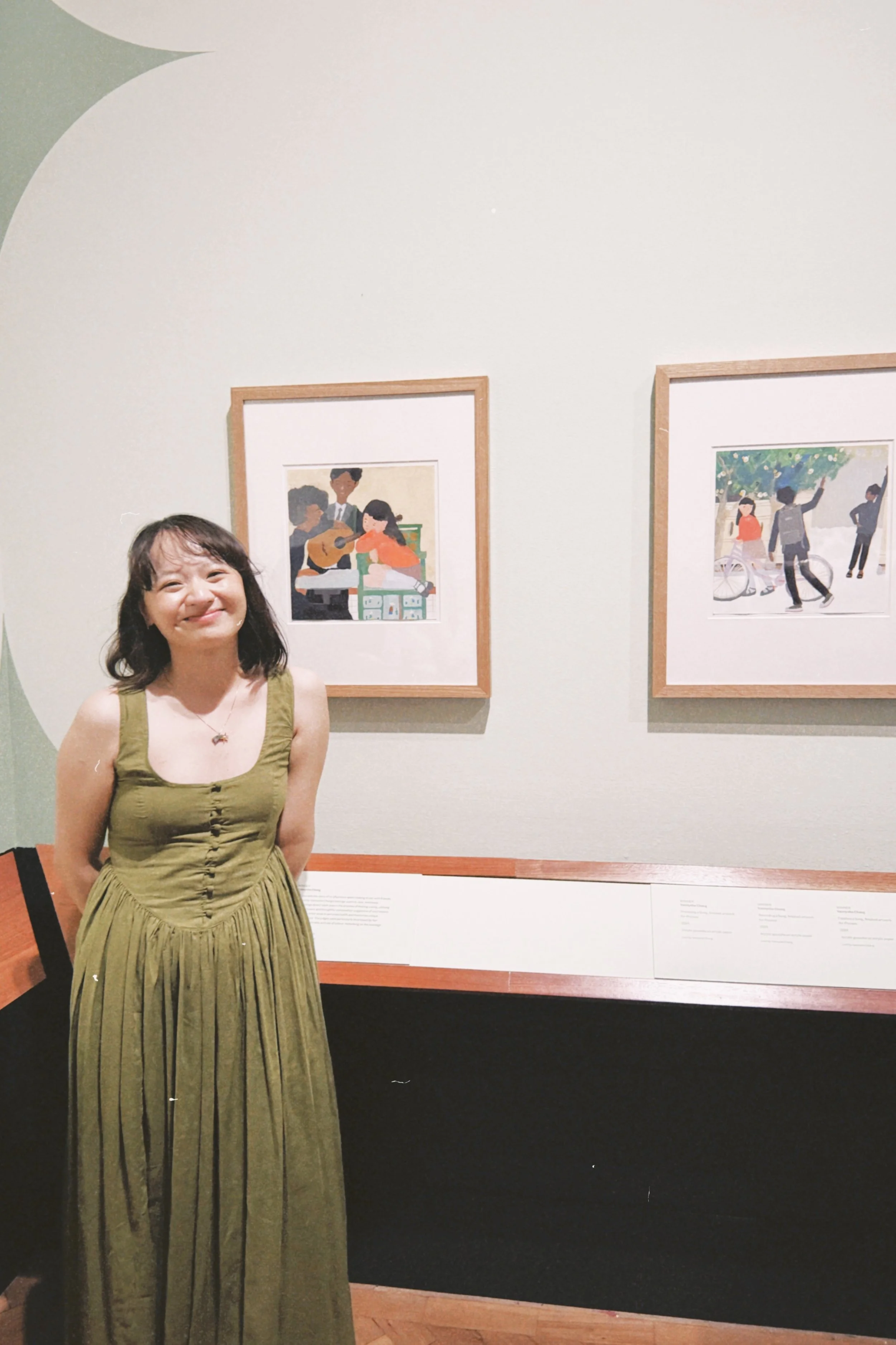 Vannysha wearing a green dress with pleats, standing in front of her work displayed at the V&A Museum in South Kensington.