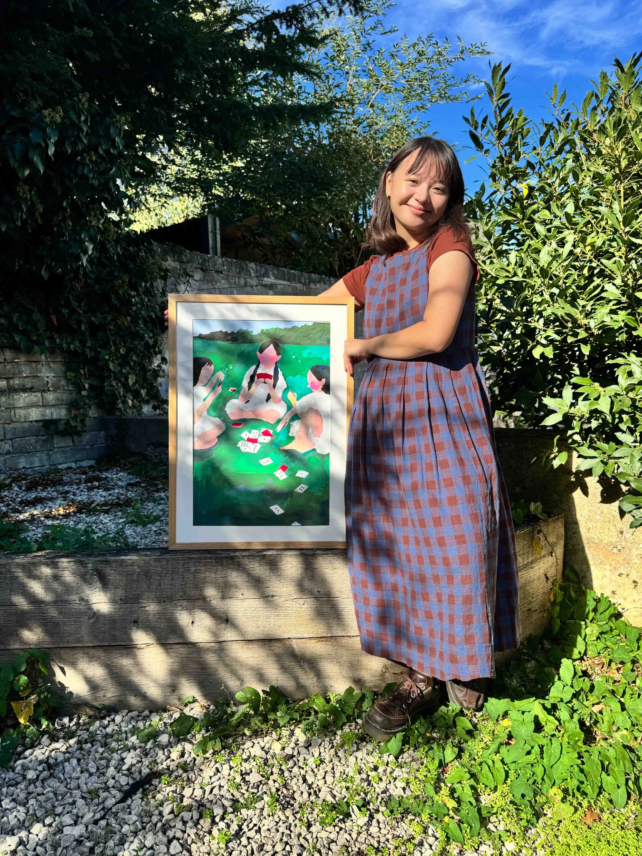 Vannysha, the illustrator, standing outside and smiling while holding her painting of three girls playing cards in a field.