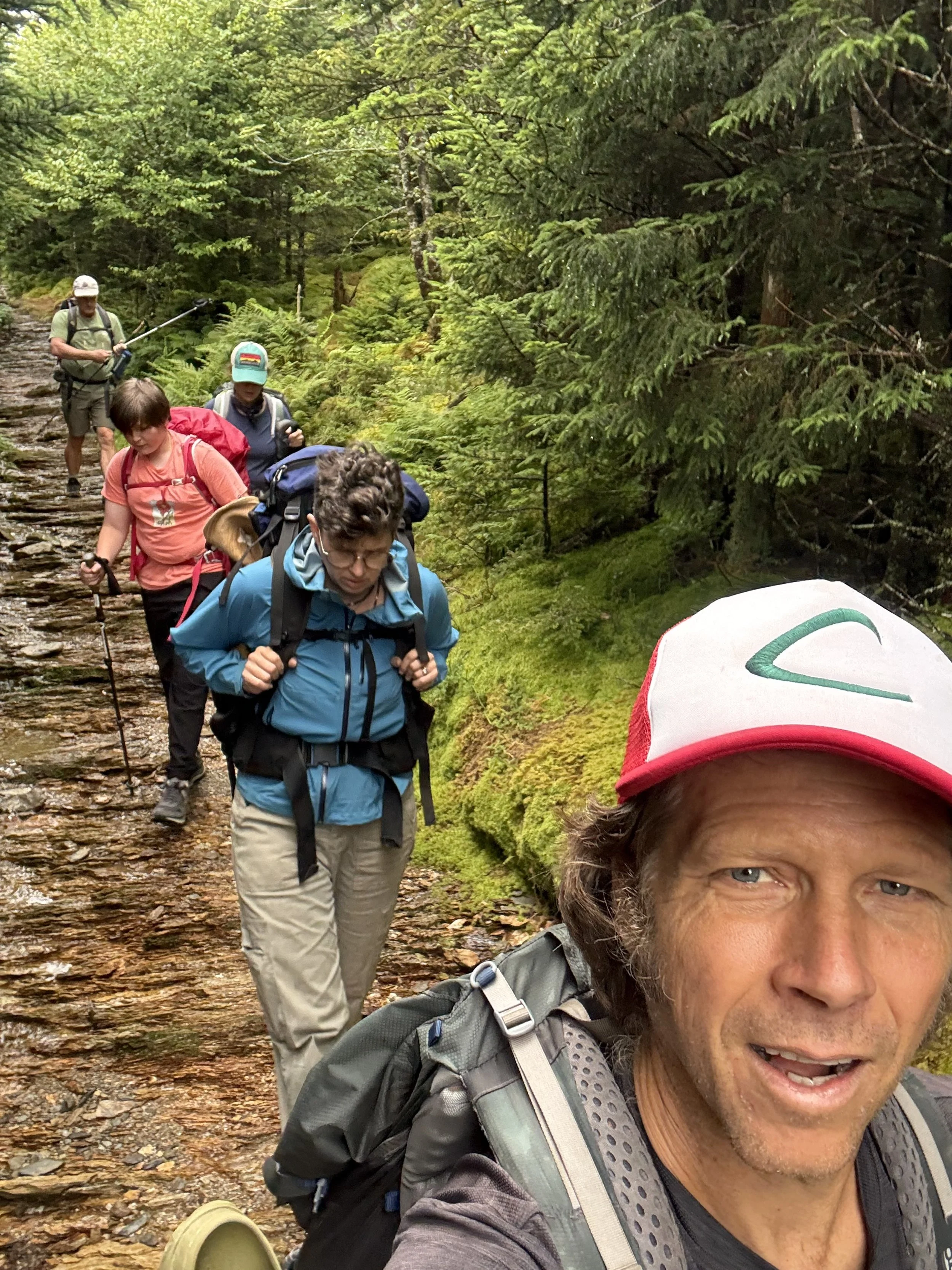 A group of hikers walking on a rocky trail in a dense green forest, with the person at the front taking a selfie.