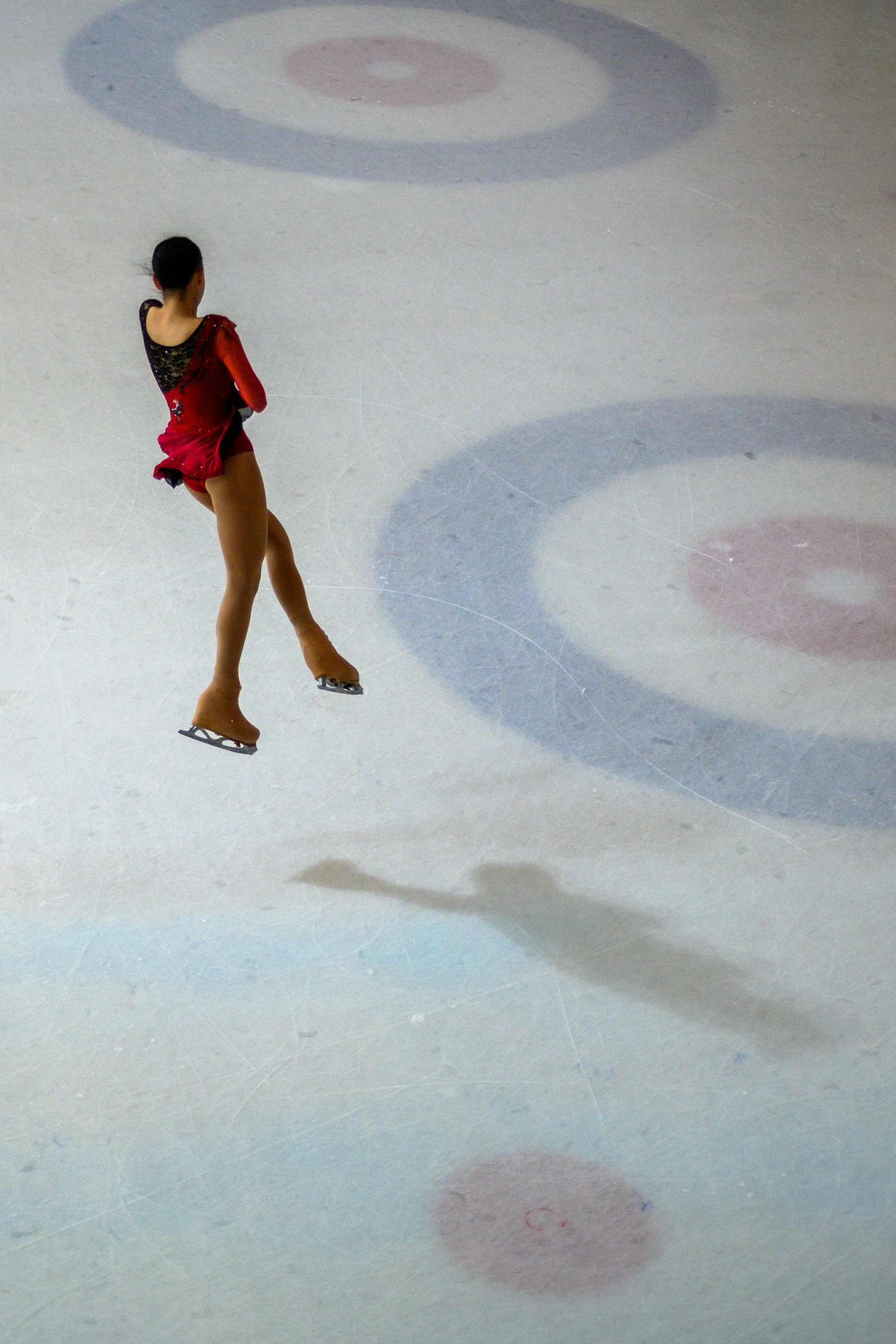 An ice skater in a red costume practicing on an ice rink with their shadow cast on the ice.