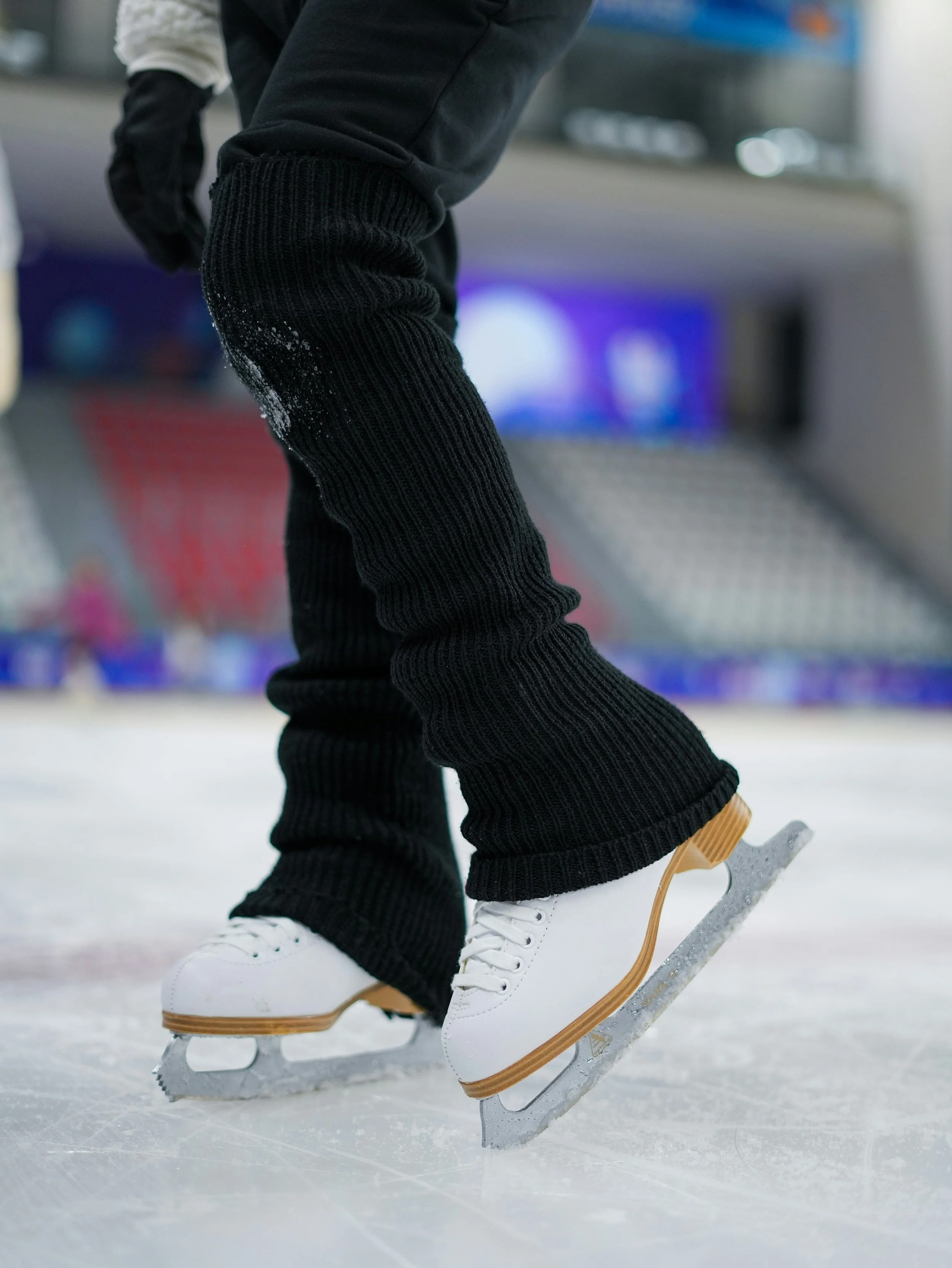Close-up of a person wearing white ice skates, black pants, and black gloves, skating on an indoor ice rink.