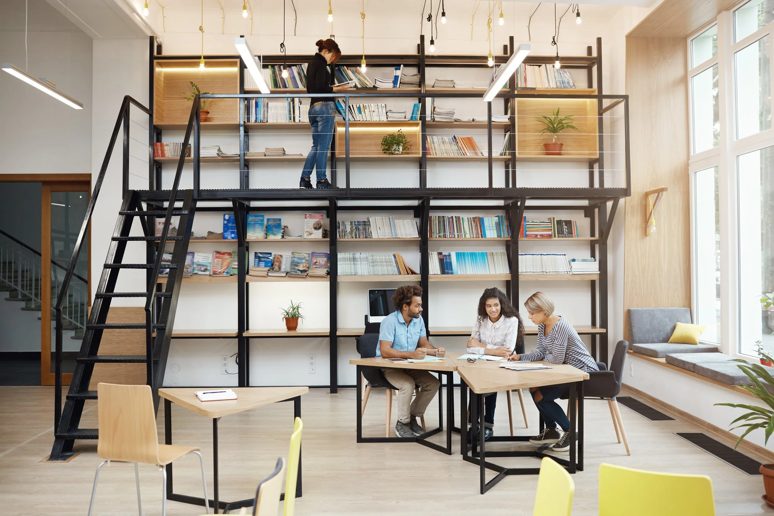 A modern coworking space with high ceilings, large windows, and a bookshelf wall. Three people are sitting together at a table working or having a meeting. A woman on a small platform level is reading a book, with a black metal ladder leading up to the platform. The space is decorated with plants and bright lighting.