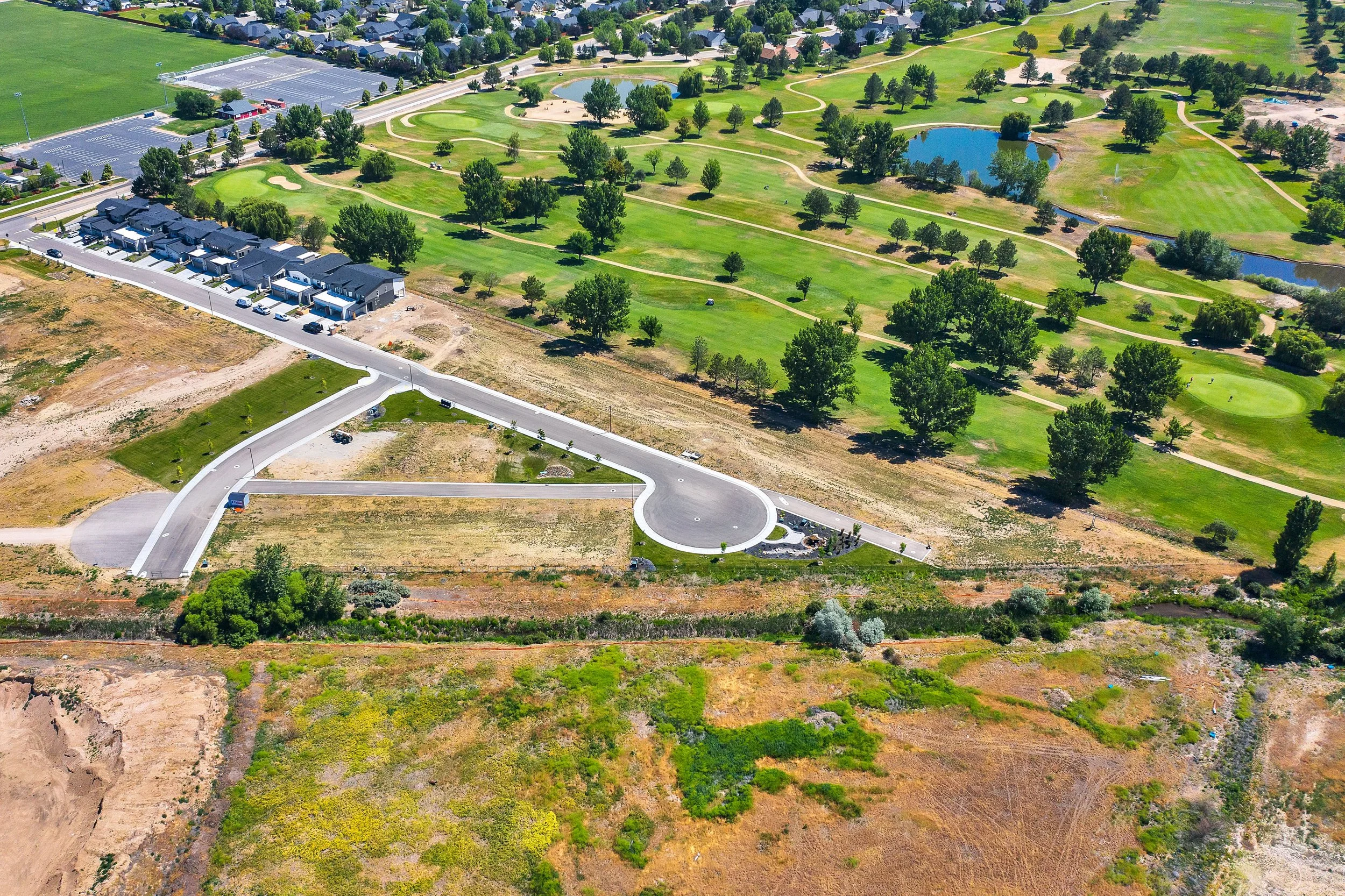 Aerial view of a golf course with green fairways, water hazards, and surrounding trees. Additionally, there is a new residential street with houses, driveway, and parked cars nearby.