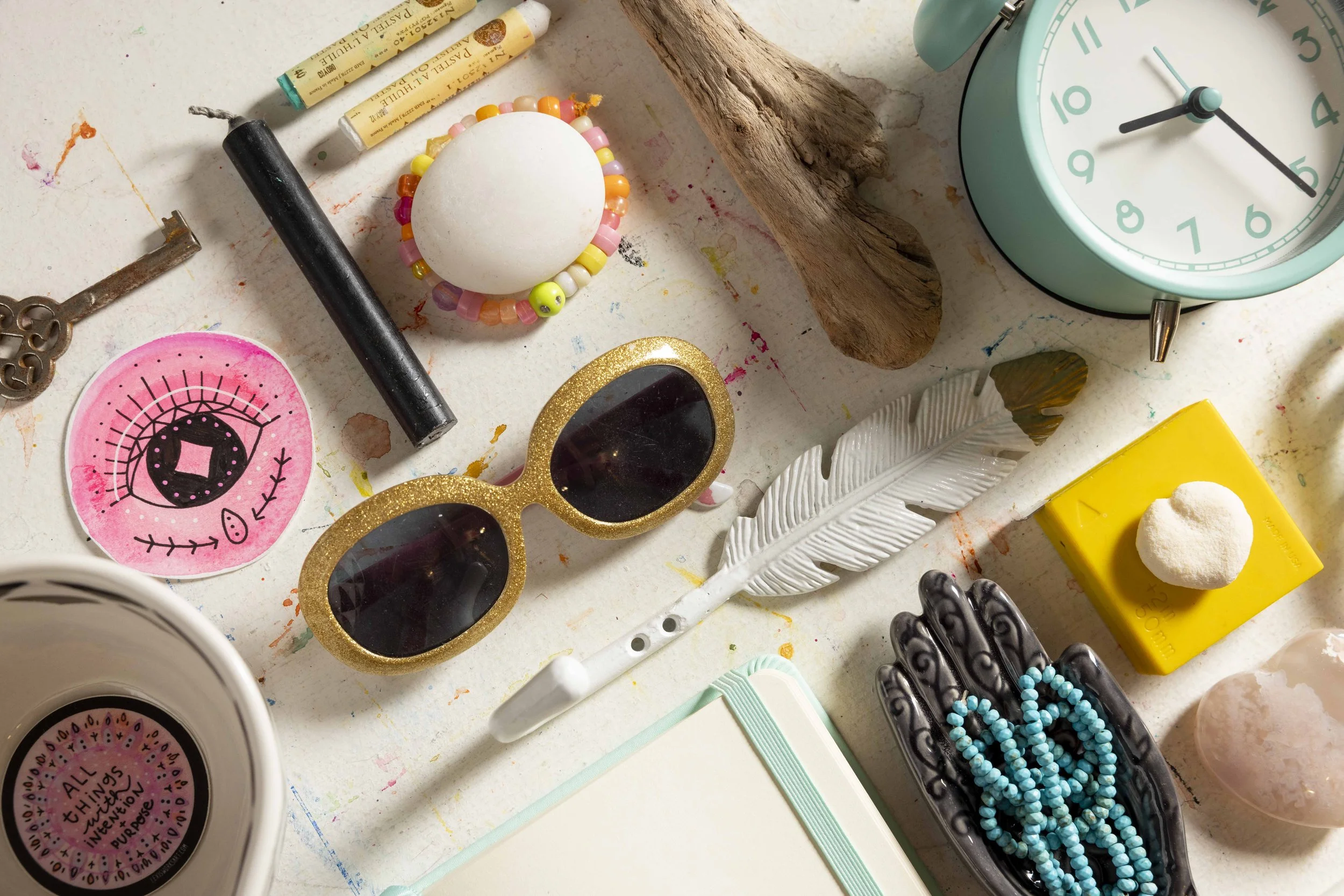 An eclectic assortment of items on a textured surface, including sunglasses, clock, beads, decorative feather, notebook, candle, key, beads, artwork, and other trinkets.