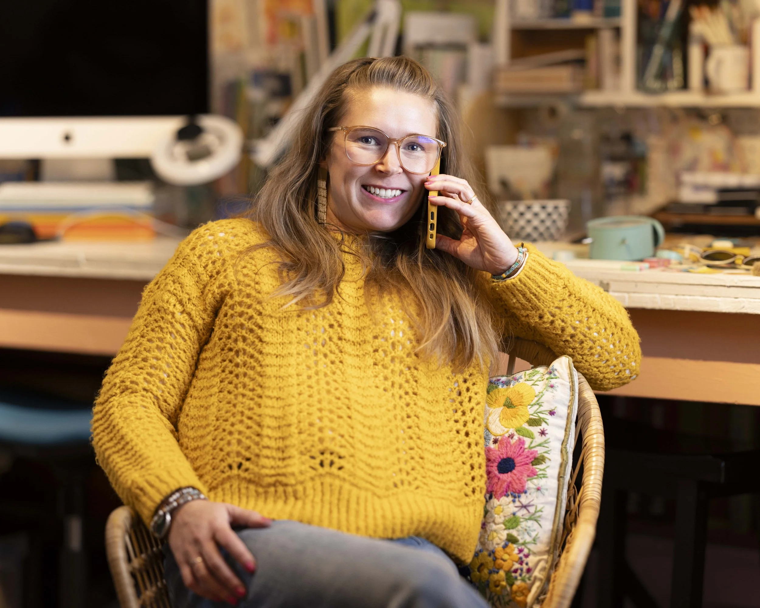 A woman wearing a yellow sweater and glasses, sitting in an office chair, holding a phone to her ear and smiling, surrounded by a desk with various items.