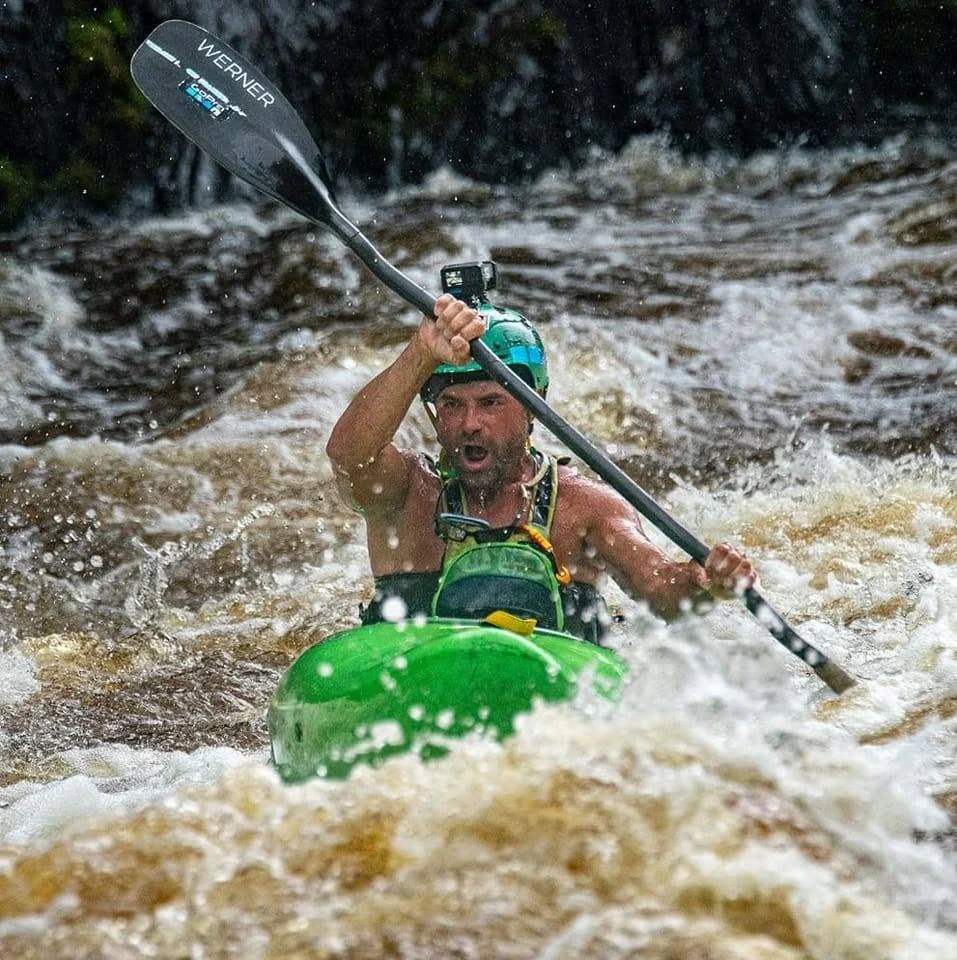 Man kayaking in white water rapids, holding a paddle above his head, wearing a helmet and life vest.